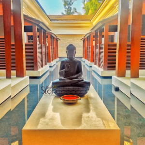 serene and elegantly composed outdoor scene featuring a dark stone Buddha statue centrally positioned at the end of a long, light-colored stone pathway or elevated platform. The Buddha is depicted in a meditative lotus position (Padmasana) with its right hand raised in the Abhaya Mudra, symbolizing fearlessness, peace, and protection, while its left hand rests gently in its lap. The statue has a calm, contemplative expression and sits upon a sculpted lotus pedestal, indicative of profound spiritual symbolism. Immediately in front of the Buddha, on the same stone platform, is a small, shallow, ornate metallic bowl, likely brass or copper, containing a vibrant reddish-orange powder, possibly kumkum or vermillion, serving as an offering. The pathway is flanked on both sides by narrow, reflective pools of dark blue water, creating a profound sense of tranquility and spaciousness. These pristine water features beautifully reflect the surrounding architecture and the bright, clear blue sky above. Beyond the water, symmetrical structures line the scene. These comprise tall, rich reddish-brown wooden pillars supporting roofs with traditional terracotta-colored tiles, and lower walls adorned with matching louvered or slatted wooden panels. These architectural elements suggest a traditional Asian or Southeast Asian style, reminiscent of a luxury resort, spa, or a temple complex, emphasizing natural materials and thoughtful design. In the distant background, under the bright sky, light-colored building facades and hints of lush green foliage, possibly palm trees, complete the peaceful vista. The lighting is bright and natural, casting subtle shadows that add depth to the scene. The overall impression is one of profound calm, spiritual contemplation, and sophisticated design, making it an ideal visual representation for articles discussing wellness retreats, meditative spaces, luxury resort architecture, spiritual gardens, or custom stone sculptures and installations for tranquil environments.