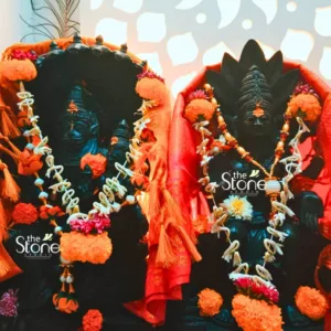 striking close-up photograph features two exquisitely carved black stone statues, likely depicting Hindu deities, adorned with vibrant offerings and textiles. On the left, a seated deity is enshrined within a deep, intricately sculpted dark stone archway, resembling a miniature temple or sacred alcove. This figure is richly decorated with multiple garlands, including delicate strings of small white flowers, possibly jasmine, and thick ropes of bright orange marigold pom-poms. A flowing, richly textured orange fabric, complete with decorative tassels, drapes elegantly over the deity's shoulders and across the top of the shrine. To the right, a second prominent black stone deity stands (or sits in a similar posture) with an elaborate, multi-tiered crown-like headdress. This figure is also profusely adorned with several garlands of white and orange flowers, prominently featuring a more elaborate beaded garland that includes a central pearlescent pendant and a small yellow flower. A luxurious red fabric, possibly silk with subtle golden embellishments, acts as a backdrop and drapes around this deity, adding to its regal appearance. Both statues are surrounded by loose clusters of fresh orange and some white/yellow flowers scattered at their base, suggesting offerings or ceremonial decorations. The background features a softly blurred, light-colored wall with decorative, leaf-shaped cutouts, which create an ethereal, halo-like effect, enhancing the sacred and devotional atmosphere. The "The Stone Studio" logo is visible on the right deity, indicating these are artisan-crafted stone sculptures. This image beautifully showcases intricate stone carving, traditional Hindu idol decoration, and the ceremonial adornment of divine figures, making it highly relevant for articles focusing on spiritual art, religious craftsmanship, temple decor, home altars, or the cultural significance of Hindu deities and their worship. The deep, rich tones of the black stone contrast beautifully with the vivid oranges, reds, and whites of the flowers and fabrics, creating a truly captivating visual representation of devotion and artistry.