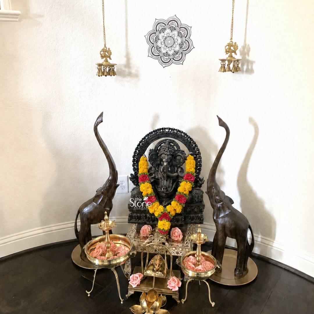 36'' Large Sitting Lord Ganapati Statue 3 A reverent home temple setup features a commanding 3-foot Black Stone Ganesha Sculpture as its centerpiece exquisitely arranged against a pristine white wall. Lord Ganesha seated majestically is adorned with a vibrant garland of yellow and red flowers highlighting the intricate carvings of the black stone. The sculpture exhibits fine details in Ganesha's form including his elephant head multiple arms and traditional attire all rendered in the deep lustrous black stone making him a focal point of devotion. Behind the Ganesha a dark ornate arch or Prabhavali further frames and accentuates the deity enhancing his divine presence. The Ganesha statue is elevated on a decorative brass stand underneath which a smaller possibly ceremonial brass swing or stand is placed holding delicate pink rose petals. Flanking the central Ganesha are a pair of tall elegant wooden elephant statues standing upright with their trunks raised symbolizing good luck and prosperity. In front of these on either side are ornate brass urulis or bowls filled with fresh pink rose petals positioned on decorative brass stands creating a symmetrical and inviting devotional space. Overhead two traditional hanging brass oil lamps with intricate designs add a classic touch though they are not lit in this image. The entire arrangement rests on a dark wooden floor contrasting with the white wall and further emphasizing the sacred space. A black and white mandala drawing or decal is affixed to the wall between the hanging lamps adding a spiritual geometric element to the backdrop. This elaborate and beautifully curated setup perfectly exemplifies the use of a Black Stone Ganesha Sculpture for Home Temple decor showcasing how a 3-foot statue can serve as the spiritual anchor in a personal sanctuary creating an atmosphere of devotion elegance and cultural richness.