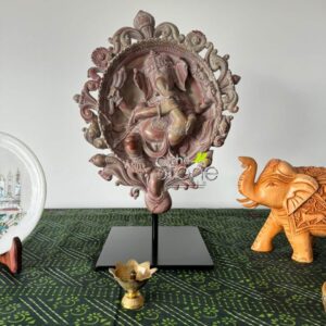 stunning, intricately carved relief sculpture of Lord Ganesha stands as the central focus of this image, presented on a modern display. The Ganesha idol is fashioned from natural, multi-toned stone, displaying a beautiful blend of earthy pink, reddish-brown, and subtle greyish veins, giving it an authentic, aged aesthetic. Lord Ganesha, the revered Hindu deity, is depicted in a comfortable, relaxed pose, seated or reclining, with his four arms holding various traditional attributes, and his trunk gently curled to hold a modak (sweet dumpling), symbolizing joy, wisdom, and prosperity. He is framed by an elaborate, circular archway, often known as a prabhāmaṇḍala, intricately carved with traditional Indian motifs including lotus petals, swirling vines, floral patterns, and what appear to be celestial beings or traditional auspicious symbols. A prominent, ornate crown-like flourish sits at the very top of the frame, adding to its regal and divine appearance. This detailed Ganesha panel rests securely on a sleek, minimalist black metal stand with a rectangular base, providing a striking contemporary contrast that beautifully highlights the stone's natural beauty and the craftsmanship of the carving. This central spiritual artwork is artfully arranged on a dark emerald green tablecloth, which features a repeating block-print style pattern of lighter green circles and diagonal lines, evoking traditional Indian textiles and adding a rich, cultural backdrop. To the left of the Ganesha sculpture, partially visible, is a white decorative plate or platter, elegantly propped on a small wooden easel. It's adorned with a detailed illustration of an architectural landmark, possibly an iconic Indian temple or palace, alongside delicate green floral inlay or painting, suggesting a finely crafted souvenir or art piece. On the right side of the frame, a charming, intricately carved wooden elephant figurine stands, its trunk raised high in a gesture often associated with good luck, welcome, and prosperity in many cultures. Its back and head are adorned with traditional patterns, showcasing fine craftsmanship and attention to detail. In the immediate foreground, directly in front of the Ganesha sculpture, a small, elegant brass diya (oil lamp) shaped like a lotus flower rests on a delicate pedestal, adding a touch of traditional ritualistic ambiance and warm golden accent. The background is a plain white or off-white wall, ensuring all the vibrant details and distinct textures of these spiritual and decorative items stand out prominently. The entire arrangement creates an inviting tableau of traditional Indian handicrafts and spiritual home decor, accentuating cultural richness, artistic craftsmanship, and the peaceful aura of devotion. This image effectively showcases a collection of unique, handcrafted decor pieces, highlighting their intricate details and cultural significance, perfect for an article discussing authentic Indian artistry, spiritual home adornments, or traditional festival decor.