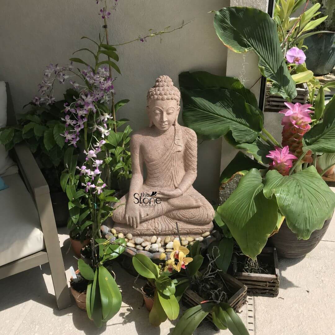 Bhumisparsha Buddha Stone Statue 3ft 3 A serene 3-foot Sandstone Buddha statue featuring an Antique Look is beautifully integrated into a lush and vibrant outdoor patio garden exuding profound peace. The statue carved from light reddish-brown sandstone depicts Buddha in a meditative seated posture specifically the full lotus position symbolizing deep concentration and spiritual enlightenment. His hands are in the Dhyana Mudra with both hands resting in his lap palms facing upwards and thumbs touching signifying meditation and inner peace. The natural earthy hue and slightly weathered texture of the sandstone give the sculpture an authentic aged and antique appearance perfectly blending with its organic surroundings. The Buddha's facial features are exquisitely carved radiating profound peace and wisdom with eyes gently closed or cast downwards in contemplation. His head is adorned with tightly coiled hair culminating in the ushnisha the cranial bump symbolizing his supreme spiritual knowledge. The flowing drapery of his robes is sculpted with soft elegant folds emphasizing the texture of the sandstone and contributing to the statue's graceful and tranquil form. The statue is seated on a base of smooth white pebbles which subtly elevates it and adds a contrasting texture. The statue is surrounded by an abundance of diverse green plants and vibrant orchids creating a rich and colorful backdrop. To the left tall purple orchids bloom while to the right large-leafed plants with pink torch ginger flowers add tropical flair. Smaller orchids with yellow and pink blossoms are arranged in the foreground. This thoughtfully designed arrangement against a light-colored wall and comfortable outdoor seating makes this 3-foot Sandstone Buddha statue an exquisite example of a garden deity with an antique charm designed to bring blessings tranquility and aesthetic beauty to any outdoor space perfectly aligning with an article focusing on such serene and timeless Buddha statues for garden decor.