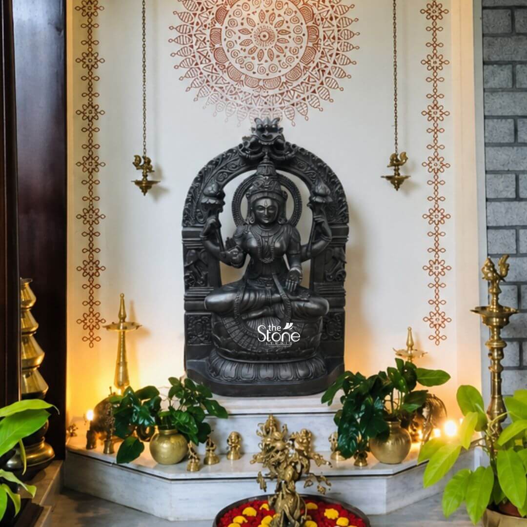 Goddess Lakshmi Statue 2.5ft 3 A full-frontal eye-level photograph capturing a beautifully adorned home temple niche dedicated to Maa Laxmi. Central to the shrine is a magnificent 2.5-foot tall blackstone Lakshmi statue meticulously hand-carved from a dark polished stone. The Goddess Laxmi is depicted seated in the padmasana upon a decorative lotus pedestal holding symbolic items in her four arms including a lotus bud and a pot of gold. Her expression is serene and she is adorned with detailed traditional jewelry and a finely carved crown all showcasing the exquisite craftsmanship of the blackstone idol. The shrine is set against a cream-colored wall flanked by two vertical patterned stencil borders. Above the Laxmi statue a large circular mandala pattern is stenciled onto the wall creating a focal point. The idol is housed within a decorative blackstone archway which is flanked by two hanging traditional brass oil lamps with lit flames. The statue rests on a white marble platform surrounded by various ritual items including two more brass diyas potted green money plants small brass elephant figurines and a decorative bowl of marigold flowers and red petals in the foreground. This 2.5-foot Blackstone Lakshmi Idol is a stunning centerpiece radiating an aura of divine grace and prosperity. Its dark textured stone contrasts beautifully with the warm golden light from the oil lamps and the vibrant greens of the plants making it a perfect addition to any temple. The overall composition with its symmetrical arrangement of traditional elements offers a sense of peace and devotion highlighting the spiritual significance and artistic value of this beautifully crafted Maa Laxmi statue.