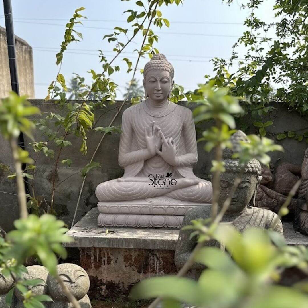 Dharmachakra Buddha Statue 4ft 3 A majestic 4-foot Brown Sandstone Buddha statue serving as an exquisite Buddha Statue for Garden Decor is peacefully situated on a stone ledge amidst a lush outdoor garden bathed in natural light. The statue carved from warm reddish-brown sandstone depicts Buddha in a serene meditative posture seated in the full lotus position symbolizing deep concentration and spiritual enlightenment. His hands are held in the Dharmachakra Mudra the gesture of teaching and turning the Wheel of Dharma with both hands at chest level right palm facing outwards and left palm inwards fingers touching to form a circle. The earthy hue of the sandstone gives the sculpture an authentic timeless elegance blending seamlessly with its organic surroundings. The Buddha's facial features are exquisitely carved radiating profound peace and wisdom with eyes gently cast downwards in contemplation. His head is adorned with tightly coiled hair culminating in the ushnisha the cranial bump symbolizing his supreme spiritual knowledge. The flowing drapery of his robes is sculpted with soft elegant folds emphasizing the texture of the sandstone and contributing to the statue's graceful and tranquil form. The statue is seated on a simple lotus-petal base which rests on a rustic stone pedestal integrating it naturally into the garden landscape. The garden setting is rich with greenery including climbing vines on the back wall and various leafy plants in the foreground some blurring gently to emphasize the main Buddha statue. Hints of other stone sculptures are visible in the blurred foreground on the right suggesting a collection or a larger sculptural garden. The sunlight casts gentle shadows enhancing the serene atmosphere. This 4-foot Brown Sandstone Buddha statue is an ideal example of a garden deity designed to bring blessings peace and aesthetic beauty to any outdoor space perfectly aligning with an article focusing on such significant Buddha statues for garden decor.