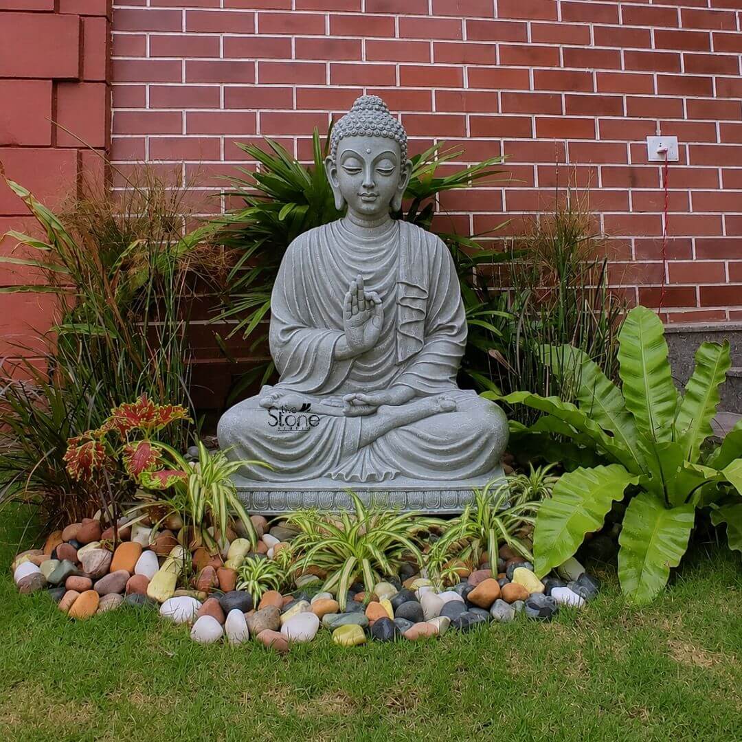 4 feet Sitting Buddha statue 3 A majestic 4-foot Fibre Buddha statue serving as an ideal Buddha Idol for Garden Decor is peacefully situated in a lush garden bed against a warm red brick wall. The statue depicts Buddha in a serene meditative posture specifically the full lotus position symbolizing deep concentration and spiritual enlightenment. His right hand is raised in the Vitarka Mudra forming a circle with the thumb and index finger while the other fingers are extended signifying the transmission of teaching and intellectual discourse. His left hand rests gently in his lap palm upwards representing contemplation and receptivity. The statue's light grey stone-like finish gives it a dignified and calming presence blending harmoniously with its natural surroundings. The Buddha's facial features are exquisitely carved radiating profound peace and wisdom with eyes gently closed or cast downwards in contemplation. His head is adorned with tightly coiled hair, culminating in the ushnisha the cranial bump symbolizing his supreme spiritual knowledge. The flowing drapery of his robes is sculpted with soft natural folds enhancing the statue's graceful and tranquil form. The statue is placed on a simple, tiered pedestal integrated into a creatively designed garden bed. The garden bed surrounding the Buddha is adorned with a variety of green plants including ferns grasses and smaller leafy plants interspersed with colorful river stones and pebbles of various sizes and hues. This vibrant arrangement creates an inviting and contemplative space perfectly suited for garden decor. The red brick wall in the background adds a touch of rustic charm and architectural interest. This 4-foot Fibre Buddha statue is an exquisite example of a garden deity designed to bring blessings peace and aesthetic beauty to any outdoor space aligning perfectly with an rticle focusing on Buddha Idols for garden decor.
