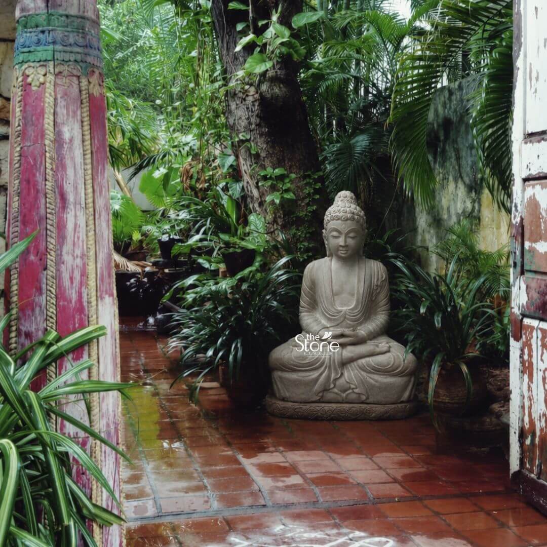 30 inch Buddha Statue 3 A tranquil 30-inch garden Buddha idol meticulously hand-carved from off-white sandstone sits in a traditional meditative pose at the heart of a secluded courtyard. The statue is positioned on a simple stone pedestal amidst a lush array of potted tropical plants including ferns and spider plants which create a natural serene backdrop. The Buddha's face is serene and contemplative with downcast eyes a gentle smile and tightly curled hair rising to a pointed ushnisha all rendered with the soft matte texture and light beige-to-grey tones characteristic of the sandstone material. The statue's robes show subtle flowing folds draped over its crossed legs and shoulders with its hands resting gracefully in the lap in the dhyana mudra of meditation. The setting for this 2.5ft Garden Buddha Idol is a rustic and inviting outdoor space. The ground is paved with reddish-brown terracotta tiles that are damp from a recent rain enhancing their color and creating subtle reflections. The statue is framed by weathered elements to the left stands a tall heavily distressed wooden pillar painted in faded shades of red and blue with visible carvings while to the right is an aged wooden door frame. Behind the statue a large tree trunk and a densely planted garden wall contribute to a feeling of peaceful enclosure as if the idol has resided there for years. Natural diffused daylight illuminates the scene highlighting the intricate carvings and the contrast between the smooth stone and the surrounding foliage. This photograph perfectly captures the integration of sacred art into a living garden environment making it an ideal illustration for an article about choosing a 2.5ft Garden Buddha Idol. The off-white sandstone material harmonizes beautifully with the natural greens of the plants and the earthy tones of the weathered wood and tiles. The overall composition evokes a sense of cal contemplation and timelessness demonstrating how such a statue can transform a simple patio or corner of a garden into a peaceful sanctuary for reflection and spiritual retreat. The detailed craftsmanship and thoughtful placement emphasize the idol's role as a focal point of peace and beauty.