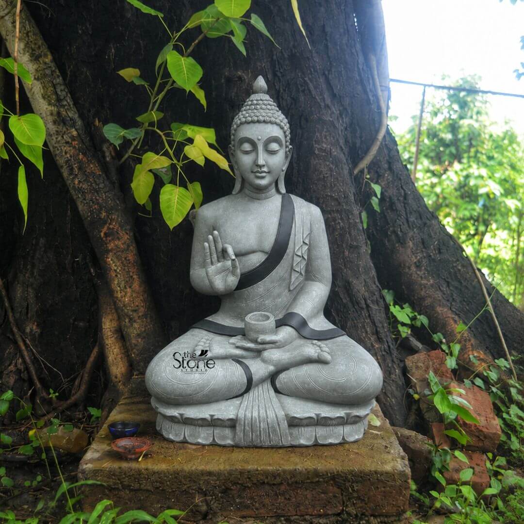 Big Buddha Statue for Outdoor 5.5ft 3 A medium shot captures a majestic five-and-a-half-foot-tall statue of the Buddha meticulously crafted from durable weather-resistant fibre. The statue is finished in a textured grey stone-like appearance perfectly suited for garden decor and is depicted in a traditional seated meditation pose known as Dhyana Mudra upon a detailed lotus pedestal. The Buddha's hands are held in a specific gesture with the right hand raised in Vitarka Mudra and the left hand resting in his lap holding a small cylindrical offering pot. His face conveys a profound sense of inner peace and serenity with eyes half-closed and a gentle compassionate expression. The Buddha figure is adorned with a clean defined black trim that outlines his robe or sanghati adding a subtle contrast to the grey body and highlighting the garment's folds. His head is covered in the characteristic small snail-shell curls topped with the ushnisha protrusion symbolizing wisdom and enlightenment. The fibre material exhibits a finely detailed texture that mimics natural carved stone giving the large idol a solid substantial presence while remaining relatively lightweight. The statue is positioned outdoors nestled at the base of a large dark-barked tree which serves as a natural and stable backdrop. In the upper left a branch of a sacred Peepal tree with its distinctive heart-shaped green leaves overhangs the scene creating a sense of sacred space. The ground around the statue is covered in natural earth and small green weeds and to the left two small weathered offering bowls one blue and one brown sit on the mossy brick plinth that raises the statue slightly. To the right a few old broken red bricks are partially buried in the foliage. Natural daylight filters through the leaves casting soft shadows that emphasize the statue's contours and rich textures making it a captivating focal point for any garden landscape.