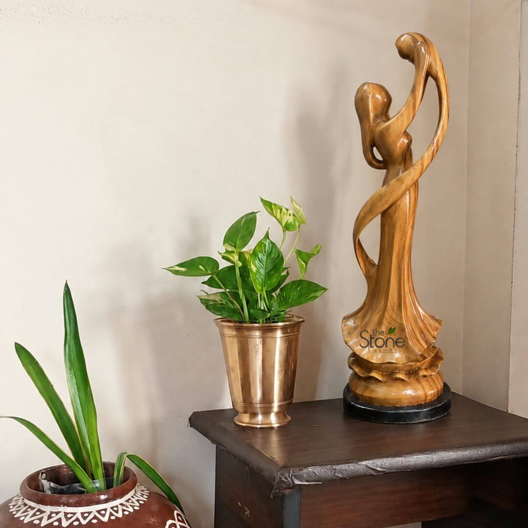 A two-and-a-half-foot-tall hand-carved wooden sculpture depicts a mother and child in a tender embrace standing gracefully on a dark wooden side table. The statue, sculpted from a single block of warm honey-toned wood showcases the natural grain and a smooth polished finish. The mother is depicted with flowing draped lines that form her garment symbolizing movement and her arms are wrapped lovingly around the child who is held securely in her embrace with their faces touching in a gesture of deep affection. The sculpture captures a timeless bond with the figures forms rendered in an abstract fluid style that emphasizes emotion over fine detail. The wooden statue is positioned on the right side of the dark wood table which has a simple rustic design. To its left sits a metallic hammered-finish brass pot holding a vibrant variegated Pothos plant with green and cream-colored leaves. The table itself is placed against a plain off-white wall. In the lower-left foreground, a larger, traditional terracotta pot with a patterned white rim holds another green plant, adding a touch of earthy texture to the scene. Natural daylight from an unseen window to the right illuminates the sculpture and plants highlighting the rich texture of the wood and the metallic sheen of the brass pot. This exquisite two-and-a-half-foot wooden mother and child statue is a perfect embodiment of maternal love and family connection, masterfully crafted to serve as a meaningful decorative centerpiece. The elegant fluid design and the warmth of the natural wood make it a versatile addition to any home decor, whether placed in a living room bedroom or nursery. The substantial size ensures it makes a visual impact, while the subject matter resonates universally making it an ideal gift for new mothers or a cherished piece for anyone celebrating the family unit. The sculpture's presence brings a sense of peace, love, and artistry to its surroundings.