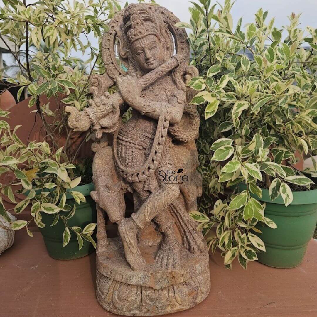 Stone Krishna with cow statue 2ft 3 A magnificent 2-foot Sandstone Krishna statue perfectly suited as a Krishna Statue for Home Decor stands gracefully on a warm reddish-brown surface likely a terracotta patio or shelf amidst lush green and white variegated plants. The statue carved from light brown sandstone depicts Lord Krishna in his iconic Tribhanga pose with a gentle sway to his body radiating charm and divine playfulness. He is exquisitely detailed with an elaborate crown intricate jewelry adorning his neck arms and ankles and finely sculpted flowing garments all emphasizing the natural texture and beauty of the sandstone. He holds a flute to his lips poised as if playing a melodious tune. The craftsmanship is evident in the delicate features of his face including his serene expression and the intricate detailing of his crown and ornaments. The flowing drapery of his garments is intricately sculpted to show texture and movement clinging gracefully to his form. A small docile cow or calf is depicted near his feet a common motif associated with Krishna symbolizing his role as a protector and caretaker. A halo-like circular aureole with ornate patterns frames his head further emphasizing his divine presence. The statue rests on a tiered round base adorned with lotus petal carvings adding to its spiritual and aesthetic appeal. The surrounding potted plants with striking green and white leaves create a vibrant and natural backdrop enhancing the statue's serene presence. This 2-foot Sandstone Krishna statue is an exquisite example of religious artistry designed to bring blessings, peace and aesthetic beauty to any home making it an ideal focal point for devotion or decorative accent aligning perfectly with an article focusing on such charming Krishna statues for home decor.