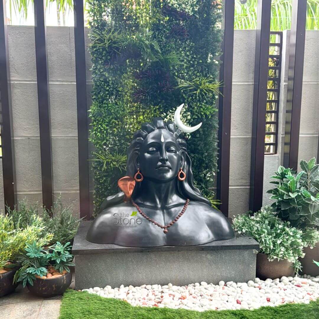 3 feet Adiyogi Statue 5 striking and highly detailed dark stone bust of Lord Shiva is prominently displayed in a serene garden setting, depicted from the chest up. Shiva's face is calm and meditative, with his eyes gently closed, and a clear white vertical mark (tilak or tripundra, signifying his third eye) adorning his forehead. His hair is styled in long, wavy dreadlocks, and a luminous white crescent moon (Chandra) is elegantly placed on the right side of his head. Around his neck, a mala of dark, reddish-brown beads, likely rudraksha, hangs gracefully. Distinctive, intricate earrings are visible: a coiled, copper-toned serpent (representing Nagakundala, often associated with Vasuki) is in his left ear, while a simpler, matching hoop is in his right ear. The sculpture, crafted from a smooth, dark material resembling polished granite or black stone, rests on a sturdy, dark grey rectangular pedestal. Behind the bust, a vibrant and lush green vertical garden wall creates a natural, verdant backdrop, featuring a variety of artificial foliage in different shades of green, interspersed with some darker, reddish-purple leaves, giving the impression of a dense, living plant wall. The vertical garden is framed by dark brown wooden slats, with a lighter grey wall visible beyond. In the foreground, a meticulously designed ground cover consists of a bed of clean, smooth white pebbles, subtly mixed with a few reddish or pinkish stones, bordering a patch of bright green artificial turf. To the left and right of the pedestal, small potted plants, some housed in unique coconut shell planters, add to the organic garden aesthetic. The overall scene is bathed in bright, natural light, creating a peaceful and spiritual ambiance. This garden sculpture exemplifies how traditional Hindu deities can be integrated into contemporary outdoor or indoor spiritual spaces, ideal for garden decor, meditative areas, or as a focal point in a wellness or home design article.