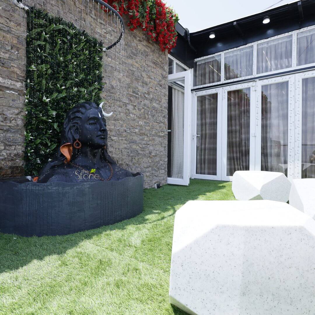 3 feet Adiyogi Statue 3 serene and richly detailed outdoor living space, likely a patio or garden, centered around a large, dark stone sculpture of Lord Shiva emerging from a rectangular basin. The sculpture, crafted from what appears to be a black or very dark grey stone, depicts the Hindu deity in a meditative pose with a crescent moon adorning his matted locks (jatamata) and water gently cascading over his head from a curved, black metallic shower-like fixture above. He wears a distinctive copper-toned earring in his right ear and a rudraksha bead mala (necklace). The subtle brand logo "The Stone" is visible on the side of the basin, indicating the source or material. Behind the Shiva fountain, a vibrant green vertical garden of lush ferns and foliage climbs a textured wall, which itself is clad in rustic, natural stone blocks in varying shades of grey and brown. Above this, from an unseen overhang, cascading vibrant red flowering plants, resembling bougainvillea, add a dramatic splash of color against the earthy tones of the stone. The ground is covered in meticulously maintained, bright green artificial turf, creating a neat and verdant carpet. Leading away from the fountain are several modern, abstractly shaped white stone benches or stools, characterized by their faceted, geometric design, offering a contemporary seating arrangement. To the right, a building with sleek white-framed glass doors and windows opens onto this outdoor area. One door is slightly ajar, hinting at seamless indoor-outdoor living. The overhead structure is dark, possibly black, with recessed lighting. The overall scene blends traditional spiritual iconography with modern landscape architecture and garden design, showcasing thoughtful use of natural and artificial stone elements, greenery, and water features to create a tranquil, sacred, yet stylish outdoor sanctuary.