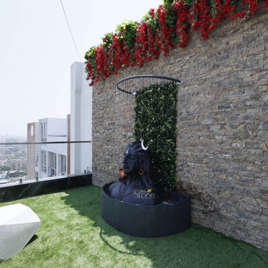 3 feet Adiyogi Statue 8 long and descriptive alt text for this image would be: "A stunning and tranquil rooftop terrace garden, designed with a blend of modern aesthetics and spiritual elements. The focal point is a large, dark grey or black stone statue of Lord Shiva, resembling the meditating Adiyogi bust, positioned centrally. Water gracefully cascades from a minimalist black circular shower ring positioned overhead, showering down onto the deity's head and collecting in a dark, semi-circular basin at its base, creating a serene water feature. The Shiva statue features intricate details including a white crescent moon (Chandra) on his forehead, long flowing dreadlocks (Jata), traditional earrings, and mala beads around his neck, all rendered in a smooth, dark stone finish. Behind the statue, a lush green vertical garden panel provides a vibrant, naturalistic backdrop against an impressive feature wall. This wall is constructed from horizontally laid, multi-toned natural stacked stone veneer, showcasing a rich texture and earthy hues. Above the stone wall and vertical garden, a vibrant cascade of artificial red flowers, resembling bougainvillea, mixed with green foliage, drapes gracefully, adding a striking splash of color and softness to the hardscaping. The entire terrace floor is covered with bright, well-maintained artificial green turf, suggesting an inviting and low-maintenance outdoor living space. To the far left, a glimpse of a modern, geometric white stone bench or planter adds another contemporary touch. In the background, under a clear sky, a sleek glass railing separates the terrace from an expansive urban city skyline, with numerous residential and commercial buildings visible in the distance, emphasizing the elevated and luxurious setting. A subtle 'the Stone Studio' watermark is visible on the statue, indicating the likely designer or supplier of this elegant outdoor stone sculpture and landscaping. This image beautifully showcases bespoke outdoor design, blending spiritual elements like the Shiva statue with modern landscaping, stone veneer finishes, vertical gardens, and a unique water feature, ideal for articles on luxury terrace design, custom outdoor sculptures, spiritual garden concepts, or creating serene urban retreats."