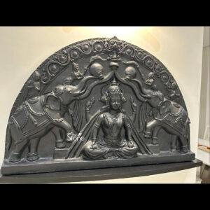 A detailed three-quarter interior photograph showcases a magnificent 3-feet tall black-patina Fibre Maa Laxmi wall mural statue mounted on a clean off-white wall in a modern living room. The semicircular or arch-shaped mural is presented from a slightly elevated angle offering a clear view of the deep intricate relief work that defines the sculpture. The deep black finish with subtle highlights catching the light mimics the appearance of antique bronze or stone adding a sense of rich history and spiritual depth to the contemporary space. The central figure is Goddess Laxmi depicted in a graceful seated posture upon an ornate lotus throne facing forward with a benevolent serene expression. She is heavily adorned with traditional temple jewelry including elaborate crowns necklaces armlets and belts all rendered in high relief. Her four arms hold symbolic attributes the upper right holds a lotus the upper left holds a pot of gold the lower right is in the gesture of blessing and the lower left holds a stack of coins symbolizing wealth and prosperity. Flanking the Goddess are two intricately carved elephants with their trunks raised in a gesture of ceremonial bathing showering her with water. The entire composition is framed by a dense deeply carved arch of floral motifs scrollwork and small divine figures demonstrating the high level of artisanal skill involved in creating this fiberglass sculpture. The mural is set against a neutral wall making it the undeniable focal point of the room visible behind a dining area and living space. The lighting from the wall sconce above the mural accentuates the three-dimensional quality and texture of the black-patina fibre making it a powerful piece of sacred art for home decor in Mumbai.