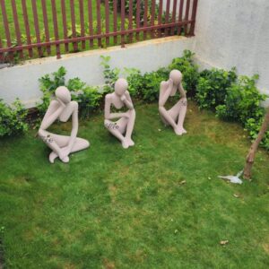 wide, high-angle shot captures three minimalist, light beige or pale pink humanoid sculptures arranged sequentially on a lush, vibrant green lawn. The figures, appearing to be crafted from smooth stone or resin, are positioned from left to right across the mid-ground. The leftmost sculpture sits in a relaxed, cross-legged pose, its head slightly tilted and its right hand gently resting against its chin, conveying a sense of calm contemplation or deep thought. The central figure is seated with knees bent and close together, forearms resting on its knees, and its head bowed with a hand supporting its face, suggesting introspection, weariness, or profound meditation. The rightmost sculpture mirrors the seated posture of the middle one but has its head completely submerged in its hands, evoking feelings of deep despair, overwhelming thought, or profound absorption. The manicured green grass provides a soft, natural base, with subtle variations in shade hinting at sunlight. In the background, a low, textured white wall runs horizontally, topped by a dark brown slatted fence. A dense line of diverse green foliage and bushes grows against the wall, adding a tranquil, natural backdrop. A thin tree trunk is visible on the far right, just beyond the last sculpture. This serene garden setting, combined with the figures' distinct pensive and reflective poses, powerfully conveys themes of mindfulness, introspection, mental well-being, and emotional states, making it an ideal visual for articles discussing self-reflection, stress relief, garden decor ideas, or contemporary outdoor sculpture.