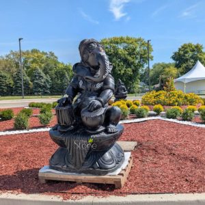 striking, large black stone statue of Lord Ganesha, the elephant-headed Hindu deity, is prominently featured in a vibrant outdoor landscaped garden on a bright, sunny day. The Ganesha sculpture, which appears to be carved from polished dark stone like granite or black marble (as suggested by "the Stone" watermark), depicts the Hindu god seated in a relaxed posture with his knees bent. He is actively playing two drum-like musical instruments – one held in each hand, resembling a tabla or mridangam set. His elephant trunk curls gracefully to the right, and his large, fan-like ears frame his face. Traditional white paint markings adorn his trunk, forehead, arms, and chest, along with a distinct red tilak (bindi) on his forehead, highlighting his divine features. The Ganesha statue rests on an ornate lotus-petal-shaped base, which itself is placed on a wooden pallet, suggesting it is either a recent installation, part of an exhibition, or awaiting final permanent placement. The statue is situated within a meticulously maintained garden bed covered in reddish-brown wood mulch. This bed is edged with neat lines of white gravel, which define sections containing small, rounded green shrubs. Beyond these shrubs, a vibrant display of brilliant yellow flowering plants, possibly marigolds, forms a lush, colorful border. Further in the background, another smaller, dark stone seated statue, possibly depicting Buddha or another meditative figure, is partially visible nestled among the yellow flowers. The background unfolds with a dense line of various green trees, including both coniferous and deciduous types, under a clear, bright blue sky dotted with a few wispy white clouds. To the far right, a large white event tent with a pointed roof is visible, hinting at a cultural celebration, festival, or gathering. The overall scene conveys a sense of spiritual peace and meticulous care in landscaping, showcasing a significant piece of religious art that likely serves as a central feature for a Hindu temple garden, cultural center, or a special outdoor event celebrating devotion and craftsmanship. The image highlights the beauty of natural stone sculptures and their role in creating sacred and inviting spaces.