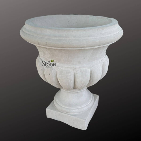 striking, large, light gray garden urn or planter, presented against a deep charcoal gray background, showcasing its elegant classical design. The vessel appears to be crafted from durable cast stone or natural aggregate, exhibiting a subtly textured, matte finish with minor natural speckling that enhances its authentic stone-like appearance. The urn features a generously wide, open top, designed to accommodate lush plantings, bordered by a substantial, multi-tiered rolled rim that adds a refined architectural touch. Below this prominent rim, the urn's elegant, bulbous body is adorned with approximately nine distinct, vertical fluted or scalloped details, creating a dynamic play of light and shadow across its surface. These graceful curves evoke timeless neoclassical or classical garden design aesthetics, ideal for a sophisticated outdoor space. The planter smoothly tapers downward from its decorated bowl to a robust, circular pedestal, which then transitions to a solid, stable square plinth base. This substantial construction suggests both aesthetic appeal and practical durability, making it suitable for heavy-duty use in various outdoor settings. Subtly visible on the upper body of the urn is a watermark-style logo for 'the Stone STUDIO', indicating its origin and quality. This timeless decorative garden feature is perfectly suited for enhancing grand gardens, patios, terraces, entryways, or as a focal point in landscape design. Its heavy-duty construction ensures longevity and resilience against the elements, making it an excellent choice for permanent botanical displays and adding a touch of classical elegance to any exterior environment.