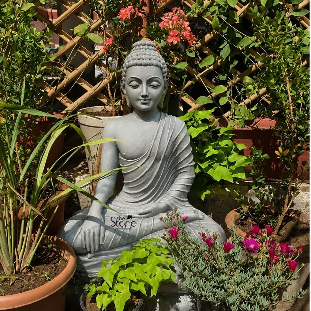 Bhumisparsha Buddha Idol 3ft 3 A tranquil 3-foot Fibre Buddha statue serving as an exquisite Fibre Buddha Idol for Home Decor is beautifully integrated into a lush and vibrant outdoor garden setting. The statue crafted from light grey fiber with a smooth finish depicts Buddha in a serene meditative posture specifically the full lotus position symbolizing deep concentration and spiritual enlightenment. His hands are in the Dhyana Mudra with both hands resting in his lap palms facing upwards and thumbs touching signifying meditation and inner peace. The light grey hue of the statue provides a calming contrast to the colorful surrounding foliage. The Buddha's facial features are finely carved radiating profound peace and wisdom with eyes gently closed or cast downwards in contemplation. A subtle serene smile graces his lips. His head is adorned with tightly coiled hair culminating in the ushnisha the cranial bump symbolizing his supreme spiritual knowledge. The flowing drapery of his robes is meticulously sculpted with soft elegant folds emphasizing the fluidity possible with fiber material and contributing to the statue's graceful and tranquil form. The smooth surface of the fiber allows for precise detailing giving the statue a refined and contemporary aesthetic. The statue is surrounded by a rich variety of green potted plants and flowering shrubs including plants with delicate pink blooms and others with small orange-red flowers creating a vibrant and natural backdrop. A bamboo trellis or fence is visible behind the plants adding a rustic architectural element. This 3-foot Fibre Buddha statue is an ideal example of home decor designed to bring blessings peace and aesthetic beauty to any indoor or outdoor living space perfectly aligning with an article focusing on such serene and contemplative Fibre Buddha Idols for home decoration.