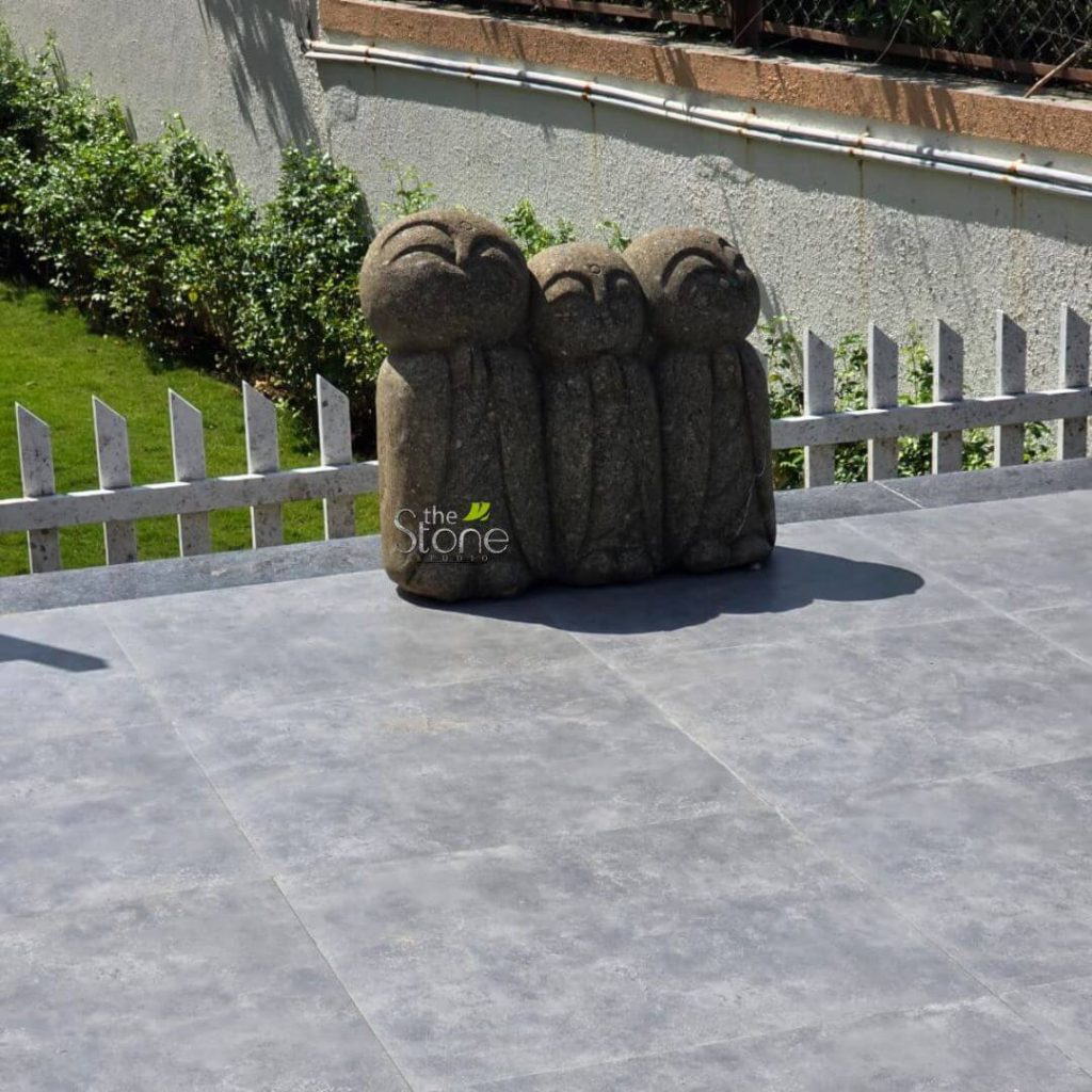 Japanese Garden Statues 28 Three charming stone Jizo statues are clustered together on a modern outdoor patio, creating a focal point of peace and simplicity. The statues, crafted from dark gray, textured stone, depict Jizo figures with simplified, rounded forms, small smiles, and closed eyes, evoking a sense of calm and contentment. They stand closely side-by-side, appearing to be in contemplation or prayer. The natural, rustic texture of the stone is evident, giving the figures an organic and weathered appearance suitable for an outdoor setting. The Jizos are placed on a spacious patio paved with large, rectangular gray tiles, which provide a contemporary and neutral backdrop. In the background, a low white picket fence separates the patio from a sloping green lawn and lush bushes, adding a touch of quaintness to the scene. Further back, a subtle metal fence and a wall with hints of greenery complete the outdoor environment. The bright natural lighting highlights the texture of the stone and the tranquil atmosphere. This placement demonstrates how Jizo statues can enhance a garden, patio, or outdoor meditation space, offering a serene presence and spiritual symbolism.
