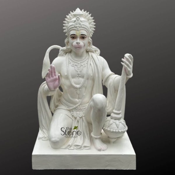 majestic, intricately carved white marble statue of Lord Hanuman, the revered Hindu deity, is prominently displayed against a minimalist dark gray background. The statue captures Hanuman in a powerful yet serene kneeling posture on a solid rectangular white base. His left knee is firmly on the ground, while his right knee is gracefully raised, conveying both humility and readiness. His right hand is raised in the 'Abhaya Mudra,' a classical gesture of blessing and dispelling fear, with the palm subtly tinted a delicate pink, suggesting vitality or a traditional ceremonial touch. In his left hand, resting on the base beside his right leg, he firmly grasps a beautifully detailed Gada (mace or club), an iconic symbol of strength, discipline, and divine authority. Hanuman's face features his characteristic monkey-like snout, with finely painted, expressive eyes that gaze forward with an alert and compassionate expression (brown pupils, black irises, white sclera, and finely arched black eyebrows). A prominent yellow 'tilak' mark, a spiritual symbol, adorns his forehead. He is adorned with an elaborate, multi-pointed crown, along with multiple strands of ornate necklaces, armlets, wristlets, and anklets, all meticulously carved from the same pristine white stone. A flowing drape of cloth is visible over his shoulder and waist, intricately sculpted to suggest movement, and his characteristic tail curls gracefully upwards and outwards behind him. The entire statue, likely a product of "The Stone Studio" as indicated by the watermark, exemplifies superior craftsmanship, showcasing smooth, polished surfaces and crisp, intricate detailing throughout the jewelry, crown, and Gada. This stunning representation of Lord Hanuman, a symbol of unwavering devotion, immense strength, and protection, is ideal for devotees seeking a focal point for worship, meditation, or as an elegant and spiritual piece of decor for homes or temples, reflecting the rich tradition of Indian marble sculpture and religious art.
