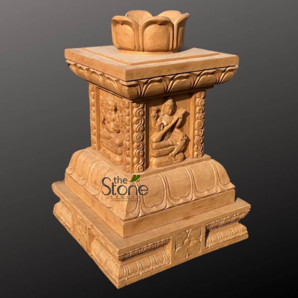 beautifully carved light brown sandstone pedestal or pillar stands prominently against a plain, dark grey background, highlighting its intricate details and earthy tones. The multi-tiered structure is rich with traditional Indian motifs and religious iconography, showcasing exceptional craftsmanship. At the very top, a prominent, closed lotus bud finial crowns a square plinth, symbolizing purity and spiritual awakening. Below this, the main rectangular body of the pillar is adorned with high-relief carvings on each of its visible sides. On the right visible panel, the Hindu Goddess Saraswati is elegantly depicted seated, playing a veena (a classical Indian stringed instrument), signifying her role as the deity of knowledge, music, art, and wisdom. To her left, another deity is carved, seated in a meditative or reverent posture, possibly Brahma or a revered sage, further enriching the spiritual narrative of the piece. These deity panels are separated by decorative vertical bands featuring intricate scrolling floral and leaf patterns, adding to the column's ornate character. The upper section of the base directly beneath the deities features a continuous frieze of stylized lotus petals, creating a harmonious transition. The pedestal then widens into a substantial, multi-layered square base, which is also intricately carved. The top edge of this lower base repeats the classic lotus petal motif, while the front panel displays a larger, central stylized lotus flower surrounded by geometric patterns and smaller floral elements, emphasizing the sacred theme. The entire stone carving, rendered in warm, natural sandstone with visible grain and texture, demonstrates deep artistic skill and a commitment to traditional Hindu artistic style. The strong studio lighting highlights the deep shadows and three-dimensional relief of the carvings, emphasizing the detailed artistry. The logo "The Stone Studio" with a green leaf emblem is subtly visible on the lower front, indicating the source of this exquisite spiritual art piece, which could serve as a focal point in a home shrine, temple, or as a sophisticated cultural decor accent.