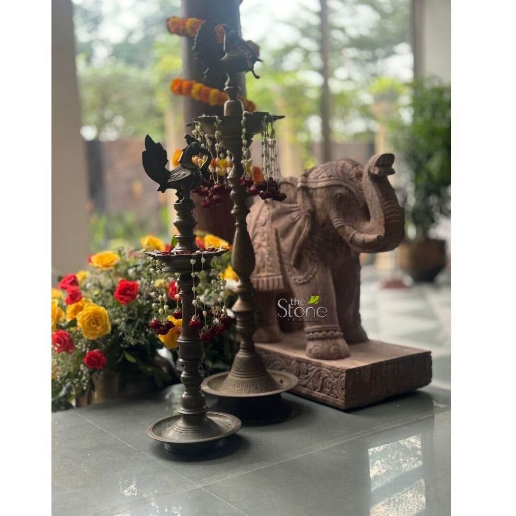 A beautifully arranged vignette showcases traditional Indian decorative elements, with a focus on a majestic 2-foot stone elephant statue. The elephant, crafted from a warm reddish-brown stone, stands prominently on a rectangular base in the mid-ground, facing to the right with its trunk raised in a welcoming gesture. Its body is intricately carved with traditional Indian motifs, suggesting ornate ceremonial attire. In the foreground, positioned slightly to the left, are two antique-style brass oil lamps (diyas), adorned with delicate strings of white flowers and small red tassels, adding a touch of festive elegance. The taller lamp has multiple arms for wicks, while the shorter one features a peacock figure at its top. To the far left, a vibrant bouquet of fresh red and yellow roses provides a lively splash of color, enhancing the inviting atmosphere. The objects are displayed on a dark gray or black countertop or table. The background is softly blurred, revealing an indoor setting with natural light filtering through a large window, hinting at a spacious and tastefully decorated home or entryway. The overall composition suggests a welcoming traditional Indian aesthetic, highlighting how such a statue can be a central decorative piece.