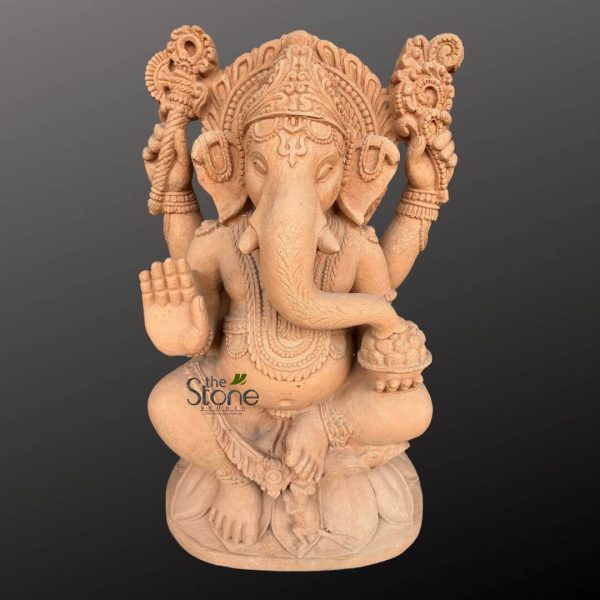 highly detailed, earthy-toned stone sculpture depicting the Hindu deity Ganesha, beautifully carved from what appears to be sandstone or a similar natural, reddish-brown material. The idol is presented against a plain, dark gray background, which makes its intricate details stand out prominently. Ganesha is shown seated gracefully in the traditional Padmasana (lotus position) atop a double-layered lotus pedestal (Padma Peetha). He has his characteristic elephant head with large, expressive ears and a gently curved trunk. A single, prominent tusk (Ekadanta) is visible, a distinctive feature of his iconography. His forehead displays intricate markings, possibly a tilak or third eye, beneath a meticulously carved, layered crown or headdress adorned with decorative motifs. The deity is depicted with four arms, symbolizing his divine power and various attributes. His upper right hand holds an ankusha (goad), while his upper left hand grasps a pasha (noose), both traditionally symbolizing guidance and control over desires. His lower right hand rests gently on a bowl of modaks (sweet dumplings), representing abundance and rewards for devotees. The lower left hand is raised in Abhaya Mudra, a gesture of fearlessness, blessing, and reassurance. The entire sculpture is exquisitely adorned with intricate carvings that mimic traditional Indian jewelry, including multiple strands of necklaces, elaborate armlets, bracelets, and anklets. Detailed drapery, folds of clothing, and a sacred thread (yagnopaveetam) across his chest are also finely sculpted, adding to the statue's rich visual texture. The craftsmanship highlights fine artisan skill, making this a significant piece of traditional Indian art or spiritual decor. The watermark "the Stone STUDIO" is subtly visible in the lower left corner. This image perfectly conveys the craftsmanship and aesthetic of a high-quality Ganesha idol, ideal for home temples, meditation spaces, or as an auspicious symbol of wisdom, prosperity, and the removal of obstacles.