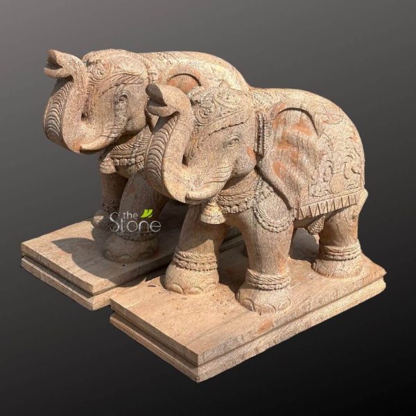 striking pair of hand-carved stone elephant statues, fashioned from light-brown or beige natural sandstone, stand majestically against a muted, dark grey to black background, creating a dramatic contrast that highlights their intricate details. The elephant in the foreground, positioned slightly to the right, showcases exceptional craftsmanship. Its trunk is elegantly raised and curled upwards, while its ears are large and textured. The entire figure is adorned with traditional, ornate carvings: an elaborate ceremonial headdress graces its forehead, intricate beaded necklaces with delicate bells hang around its neck, and a richly patterned saddle blanket or caparison covers its back, featuring detailed motifs reminiscent of traditional Indian or Southeast Asian artistry, including floral patterns and swirling designs. Decorative anklets encircle its strong legs, adding to its regal appearance. Behind and slightly to the left, the second elephant statue is visible, mirroring the first in its detailed carvings and posture, affirming it as a perfectly matched set. Both monumental elephant sculptures are securely mounted on multi-tiered, rectangular bases crafted from the same natural stone, adding to their stability and presenting them as grand, ready-to-display pieces. The textured, somewhat weathered surface of the sandstone lends an authentic, antique aesthetic to these decorative sculptures, showcasing the meticulous handiwork involved in their creation. These exquisite stone elephants are ideal as statement pieces for gardens, entryways, or large indoor spaces, reflecting cultural heritage and timeless artistry. The 'the Stone' watermark indicates the source of these high-quality, handcrafted stone art pieces, perfect for those seeking unique garden decor, traditional sculptures, or culturally inspired home accents.