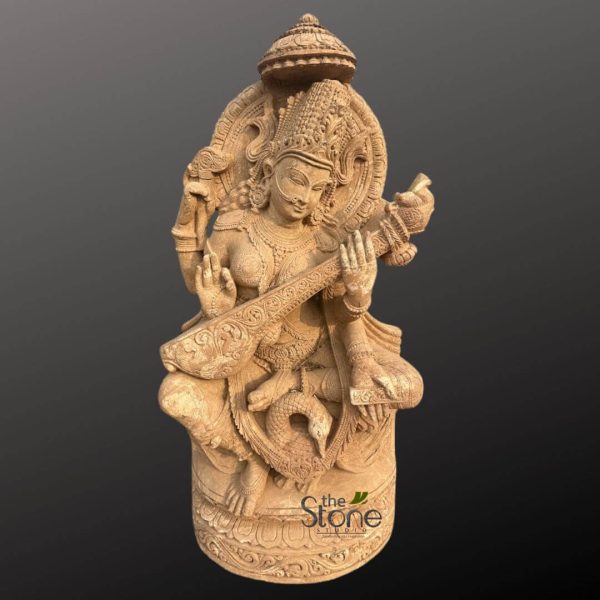 magnificent and intricately carved stone statue of the Hindu Goddess Saraswati is presented centrally against a plain, dark gray background, making the lighter-colored sculpture stand out prominently. Fashioned from a light brown or sandy-beige material, likely sandstone or a similar earthy stone, the sculpture showcases exceptional traditional Indian craftsmanship and fine detailing. Saraswati, the revered goddess of knowledge, music, art, wisdom, and learning, is depicted in a graceful, seated posture, possibly Lalitasana, on an ornate, multi-tiered circular pedestal. Her two principal hands are elegantly poised to play a highly detailed Veena, a stringed musical instrument, with her fingers delicately resting on its ornate neck and body, as if producing a divine melody. She possesses four arms, a common representation for Hindu deities. In her upper right hand, she holds an Akshamala (rosary beads), symbolizing meditation, spirituality, and the eternal cycle. Her upper left hand cradles a Pustaka (a palm-leaf manuscript or book), representing sacred scriptures, knowledge, and academic pursuits. Saraswati's serene facial expression, adorned with a delicate nose ring, conveys profound wisdom and grace. She wears an elaborate crown (mukuta) and intricate jewelry, including multiple necklaces, armlets, wristlets, and earrings, all meticulously carved into the stone. Behind her head, an ornate Prabhavali or halo, richly carved with detailed floral and geometric patterns, frames her divine presence. Her flowing drapery is subtly rendered with delicate folds, adding to the statue's lifelike quality. At the base of the statue, beneath her feet, her traditional Vahana (mount), a beautifully carved Hamsa (swan), is depicted. The swan symbolizes purity, discernment, and the ability to distinguish between good and evil, further emphasizing Saraswati's attributes. The entire sculpture rests on a circular pedestal featuring exquisite relief carvings of scrolls and patterns around its circumference, adding another layer of artistic intricacy. A discreet watermark, "the Stone Studio," is visible at the bottom right of the image, indicating the source or creator of this exceptional piece of devotional art. This detailed Saraswati murti or idol is a prime example of high-quality stone carving and traditional Indian sculpture, making it a perfect feature for articles discussing Hindu deities, spiritual art, Indian craftsmanship, or cultural decor for spaces dedicated to learning and the arts.