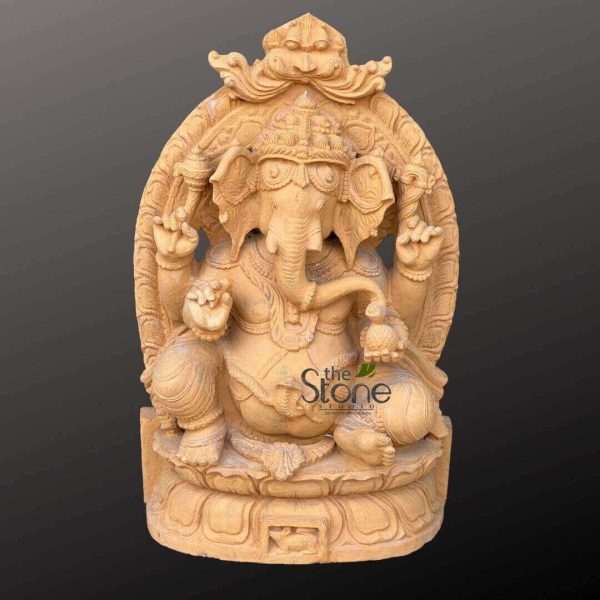 magnificent, intricately carved sandstone sculpture of the Hindu deity Lord Ganesha, presented in a full-view shot against a plain, dark gray background that accentuates its warm, light yellowish-brown tone and textured surface. This detailed stone sculpture depicts the elephant-headed god seated in a relaxed, regal posture, possibly lalitasana or royal ease, with one leg slightly elevated. Ganesha's large, benevolent elephant head features prominent, fanned ears, small, focused eyes, and a trunk gracefully curled to his left, reaching towards a modak (sweet dumpling) held in his lower left hand. A single, intact tusk is clearly visible, while the other is broken, a key iconic detail symbolizing wisdom and sacrifice. He wears an ornate, multi-tiered crown (mukut) adorned with delicate patterns and sits with a prominent pot belly, symbolizing prosperity and abundance. The deity is four-armed, characteristic of traditional Hindu iconography. His upper right hand holds an ankusha (goad), and his upper left hand grasps a pasha (noose). His lower right hand is raised in a gesture of blessing or fearlessness (abhaya mudra), offering reassurance and protection. Throughout the sculpture, Ganesha is adorned with meticulously carved jewelry, including necklaces, armlets, bracelets, and anklets, all rendered in the stone. He is seated comfortably upon a double-tiered lotus pedestal (padmasana), an emblem of purity and spiritual awakening, which rests on a simpler, rectangular base. Behind him, an exquisite, arch-shaped prabhavali (aura or frame) rises, intricately carved with flowing floral and possibly mythical creature motifs, culminating in a stylized Kirtimukha (face of glory) at its apex. This hand-carved piece showcases exceptional craftsmanship, ideal for articles discussing traditional Indian stone carving, Hindu religious art, spiritual symbolism, Vastu, home decor, or the significance of Lord Ganesha as the remover of obstacles and god of wisdom and prosperity.