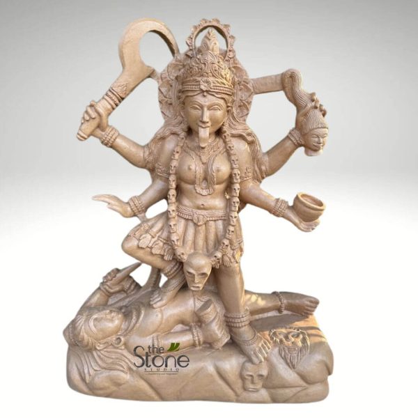 meticulously detailed, light brown stone carving of the Hindu goddess Kali, depicted in a powerful and dynamic dancing posture. She stands with her right foot firmly planted on the chest of a serene, supine male figure, traditionally identified as Shiva, who lies calmly on his back with his eyes open, looking upwards. Kali's left leg is bent, conveying movement and strength. The goddess is shown with four arms, each holding a significant attribute. In her upper right hand, she wields a large, crescent-shaped khadga or scythe, symbolizing destruction of ignorance and negativity. Her upper left hand grasps the disembodied, flowing-haired head of a demon, signifying victory over evil. Her lower right hand holds a kapala, a skull cup, often associated with offerings or drinking the blood of demons. Her lower left hand is extended in a gesture (mudra). Kali's face is fierce and awe-inspiring, with wide, intense eyes, an elaborate crown, and a long, prominent tongue extended. She wears a striking mala (garland) made of intricately carved skulls around her neck and waist, symbolizing liberation from fear and the cycle of death. Additional adornments include bangles, armlets, and a short, pleated skirt-like garment. The sculpture is carved from a light, earthy-toned stone, possibly sandstone or a similar composite, showcasing exquisite craftsmanship and fine textural details. The base of the statue is robust, featuring additional carved skulls and the face of a lion, adding to the macabre and powerful iconography. The background is a clean, neutral grey, enhancing the focus on the goddess figure. A discreet "The Stone Studio" watermark is visible on the lower front of the base, indicating the origin or vendor of this piece of devotional Hindu art. This intricate piece exemplifies traditional Indian religious sculpture, embodying themes of divine power, destruction of evil, and liberation.