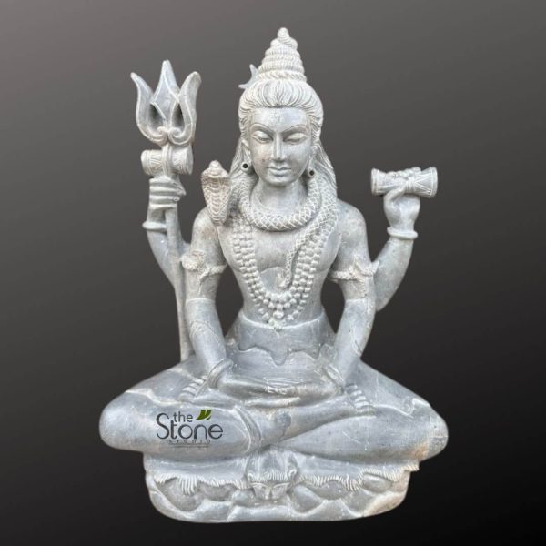 meticulously carved dark grey stone statue of Lord Shiva, seated in a serene meditative posture (Padmasana) on a lotus pedestal, fills the frame against a plain, deep charcoal grey background. The sculpture, rendered with fine detail, depicts Shiva with four arms. His upper left hand gracefully holds a finely sculpted Trishul (trident), while his upper right hand holds a Damaru (a small hourglass-shaped drum). His lower two hands rest gently in his lap, palms upward, likely in Dhyana Mudra, signifying deep meditation and wisdom. Shiva's face conveys a peaceful, contemplative expression, with eyes half-closed in spiritual absorption. His hair is styled in a towering Jatamukuta (matted hair topknot), with loose strands flowing down his shoulders, subtly adorned. He wears simple round Kundala earrings. A prominent Naga (cobra serpent), with intricately carved scales, coils around his neck and drapes over his left shoulder, its hood slightly raised. Multiple strands of beaded necklaces (Malas) adorn his chest, along with armlets and wristlets carved into the stone, highlighting the body's musculature and grace. He is depicted wearing a traditional dhoti with gentle folds, suggesting movement and drapery. The uniform dark grey color of the stone, possibly soapstone or a similar fine-grained rock, gives the deity a timeless, ethereal quality, emphasizing the form and texture over color. A subtle white watermark for "the Stone Studio" is visible on the lower front portion of the statue. This devotional art piece beautifully captures the iconic symbolism and spiritual tranquility associated with Lord Shiva in Hinduism, making it a compelling visual for articles on Hindu mythology, sacred sculptures, or the art of stone carving.