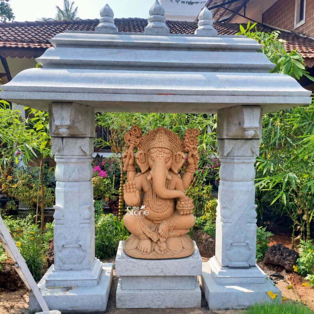 2.5 feet Lord Ganesha Statue 3 A majestic 2.5-foot Sandstone Ganesha statue perfectly suited as a Stone Ganesha for Outdoors is enshrined within a grand light-grey stone mandapam in a lush garden setting. Lord Ganesha is depicted in a seated posture crafted from warm reddish-brown sandstone which contrasts beautifully with the cooler tones of the mandapam. His iconic elephant head adorned with an elaborate crown and his four arms are intricately carved holding his traditional attributes such as a modak and an axe or goad with one hand raised in a gesture of blessing. The mandapam itself is a testament to traditional stone craftsmanship featuring four sturdy pillars with detailed capitals and bases supporting a multi-tiered roof adorned with small finials. The light grey stone of the pavilion provides a protective and reverent enclosure for the Ganesha creating a designated sacred space within the garden. The intricate carvings on the sandstone Ganesha including his jewelry sacred thread and the patterns on his halo-like structure are clearly visible showcasing the meticulous artistry involved. The entire structure is set amidst a vibrant outdoor garden with various green plants and colorful flowers flourishing around it. The natural environment enhances the spiritual aura of the Ganesha making it a focal point for devotion and aesthetic appreciation. In the background hints of traditional roof tiles and more greenery suggest a peaceful perhaps rural or suburban landscape. This 2.5-foot Sandstone Ganesha statue within its stone mandapam is an exquisite example of an outdoor deity designed to bring blessings, tranquility and a touch of traditional Indian architectural beauty to any garden or open-air space aligning perfectly with the article's focus on Stone Ganeshas for outdoor placement.