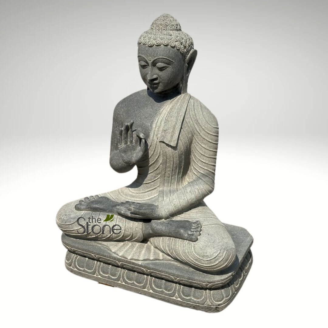 Black Granite Buddha Statue 2.5ft 6 detailed, full-body statue of Buddha, hand-carved from dark grey, finely textured natural stone, is presented against a clean, softly lit white background, suggesting a studio or product display setting. The Buddha is depicted seated in the traditional meditative lotus position (Padmasana), radiating a profound sense of peace and serenity. His eyes are gently downcast, conveying a tranquil, introspective state, with a subtle, compassionate smile gracing his lips. Intricate details include the tightly coiled hair (ushnisha) atop his head, forming a cranial protuberance, and his elongated earlobes, characteristic features of Buddhist iconography. The right hand is raised to chest level with the palm facing outwards, and the thumb and index finger lightly touching in the Vitarka Mudra, a significant gesture symbolizing teaching, discussion, and the transmission of Buddhist dharma. The left hand rests open in his lap, inviting contemplation and receptivity. His monastic robe drapes gracefully over his left shoulder and across his body, with finely sculpted, realistic folds that convey movement and softness, contrasting with the exposed right shoulder. The Buddha sits upon a smooth, round cushion-like base, which in turn rests on a sturdy rectangular plinth adorned with beautifully carved lotus petal motifs along its lower edge, symbolizing purity and spiritual awakening. The varied tones of the grey stone, ranging from a lighter, almost silvery grey in the draped fabric to a deeper, mottled charcoal in exposed areas, emphasize the natural beauty of the material and the artisanal skill involved in creating this robust, spiritual sculpture. The logo "the Stone" is subtly visible on the lower left portion of the statue, indicating its origin from a specialized stone carving or home decor brand. This exquisite piece would be an excellent visual for articles on spiritual wellness, mindfulness practices, Zen garden aesthetics, Asian home decor, outdoor meditation spaces, or the intricate art of hand-carved natural stone sculptures.