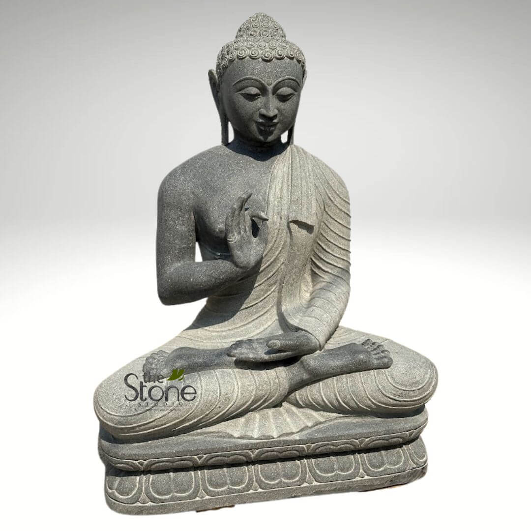 Black Granite Buddha Statue 2.5ft 5 magnificent, hand-carved dark grey granite Buddha statue is depicted in a serene meditative pose, seated in the full cross-legged Padmasana (lotus position) upon a multi-tiered, intricately sculpted lotus pedestal. The statue is made of natural, speckled dark grey stone, giving it a textural, organic feel that suggests durability and timelessness. Buddha's expression is profoundly peaceful and contemplative, with his eyes gently downcast and half-closed, signifying deep introspection and inner calm. His hair is meticulously carved into tight, circular curls, culminating in a prominent ushnisha (a cranial bump), a traditional symbol of wisdom and spiritual awakening. Elongated earlobes, indicative of his renunciation of worldly possessions, and a subtle urna (a round mark between the eyebrows), symbolizing spiritual enlightenment, are also visible. His monastic robes, known as the sanghati, are elegantly draped over his left shoulder, leaving his right arm and chest exposed. The folds of the fabric are deeply carved and flow naturally, showcasing exceptional craftsmanship and adding to the statue's realistic yet stylized appearance. The statue displays two significant mudras (symbolic hand gestures): His right hand is raised to shoulder height, palm outwards, fingers extended upwards with the thumb and forefinger lightly touching, forming the Abhaya Mudra – a gesture of fearlessness, reassurance, and divine protection. His left hand rests gently in his lap, palm facing upwards, in the Dhyana Mudra, symbolizing meditation, concentration, and the attainment of spiritual perfection. The sturdy, multi-layered lotus base provides a stable foundation for the figure, with each petal carefully carved. The statue is presented against a clean, bright white or very light grey background, ensuring that all focus remains on the detailed stone artistry. A watermark for "the Stone Studio" is visible on the lower left side of the statue. This exquisite piece of Buddhist art would be ideal for an article discussing meditation, spiritual practices, natural stone sculpture, handcrafted garden decor, or mindful home interiors, emphasizing tranquility, craftsmanship, and ancient spiritual wisdom.