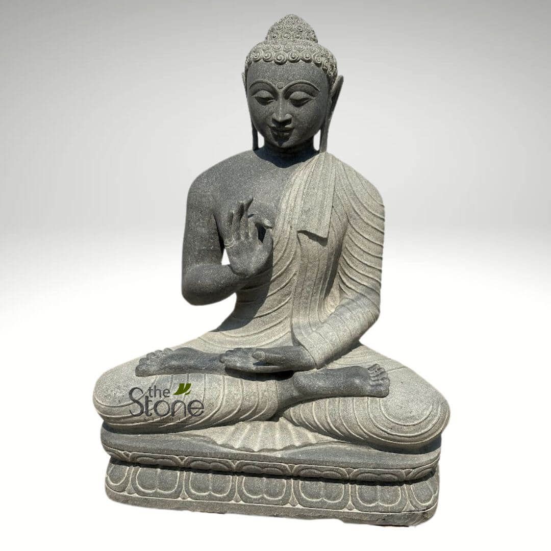 Black Granite Buddha Statue 2.5ft 4 detailed, full-body shot of a dark grey, intricately carved natural stone Buddha statue, presented against a plain, light grey to white background that accentuates its form and texture. The statue depicts Gautama Buddha seated in a serene, meditative cross-legged lotus position (Padmasana). The statue is crafted from a coarse-grained, dark grey stone, possibly granite or basalt, with subtle lighter flecks that give it a rich, earthy, and tactile appearance. The Buddha's right hand is raised to chest level with the palm facing outwards, and the thumb and index finger gently touching, forming the Vitarka Mudra, which symbolizes the transmission of teaching, wisdom, and intellectual discussion. His left hand rests open in his lap, palm facing upwards in a gesture often associated with meditation or receiving. The Buddha's face is rendered with a calm and contemplative expression, featuring half-closed, downcast eyes indicating deep meditation, a subtle smile, and elongated earlobes, traditional iconographic elements. His hair is meticulously sculpted into tight curls, culminating in an ushnisha, the raised cranial protuberance symbolizing his spiritual wisdom and enlightenment. He is gracefully draped in flowing monastic robes, carved with realistic, soft folds and pleats that suggest movement and lightness despite the heavy material. One shoulder is exposed, while the other is covered by the robe. The Buddha rests upon a rectangular pedestal, which is adorned with beautifully carved, stylized lotus petal motifs along its sides, a common symbol of purity and spiritual awakening in Buddhist art. A subtle watermark, "the Stone," is visible on the lower left portion of the base, identifying the creator or vendor. This exquisite stone carving is a perfect example of spiritual decor, ideal for creating a peaceful atmosphere in a home, garden, or meditation space, emphasizing craftsmanship, mindfulness, and the timeless serenity of Buddhist art.
