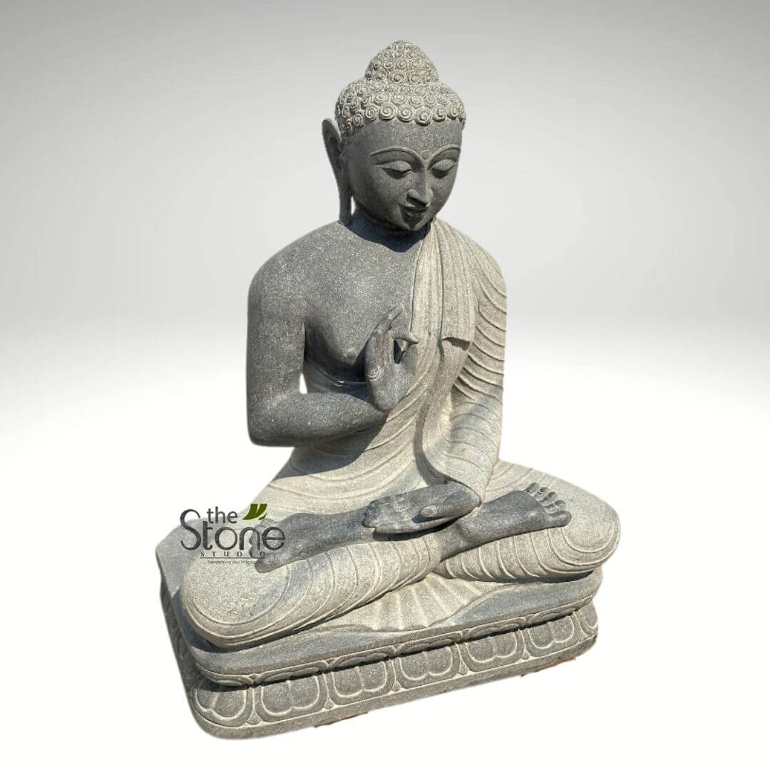 Black Granite Buddha Statue 2.5ft 2 highly detailed, handcrafted dark gray stone Buddha statue is depicted in a serene meditative state, seated in the traditional Padmasana (lotus position). Carved from a dense, speckled stone, likely granite or basalt, its texture is visible across the entire sculpture, highlighting the natural beauty and durability of the material. The Buddha’s right hand is raised to chest level in the Vitarka Mudra, where the thumb and index finger gently touch to form a circle, symbolizing teaching, intellectual discussion, and the transmission of Dharma (Buddhist teachings). His left hand rests gently in his lap, palm facing upwards in a gesture of receptivity. His face conveys deep tranquility, with eyes gently closed or downcast in an inward gaze, a serene smile, and elongated earlobes indicative of wisdom and detachment. A prominent urna (dot) is visible on his forehead, and his head is covered with tight, uniform curls leading up to the ushnisha, the cranial bump symbolizing spiritual knowledge and enlightenment. A simple, flowing monastic robe drapes gracefully over his left shoulder and across his body, with natural-looking folds that add to the sculpture's realism and flow. The statue rests on a robust square base, intricately carved with a border of traditional lotus petals along the front edge, adding a sacred and aesthetic touch. The neutral, light gray background isolates the statue, ensuring the focus remains entirely on its intricate details and contemplative presence. The visible "The Stone Studio" logo in the lower left suggests this is an exemplary piece showcasing high-quality custom stone sculpture or garden decor, blending timeless artistry with natural, durable materials, ideal for creating a peaceful and enlightened ambiance in any space. This spiritual statue embodies peace, wisdom, and mindfulness, making it a significant artisan craftsmanship piece.