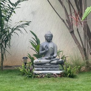 A serene 2.5-foot grey granite Buddha statue is prominently displayed in a lush outdoor garden setting, embodying tranquility and natural harmony. The Buddha is seated in a cross-legged meditative pose, with his right hand raised in the Abhaya Mudra (gesture of fearlessness and reassurance) and his left hand resting gently in his lap. His eyes are downcast, and a subtle, peaceful smile adorns his face, conveying a sense of deep calm. The statue rests on a layered base, possibly a combination of carved stone and a simple white pedestal, elevated slightly above the vibrant green grass. Surrounding the base are various green plants, including ferns and broad-leafed foliage, which naturally integrate the statue into the landscape. To the left of the Buddha, a small modern solar garden light blends into the greenery. In the background, a light-colored stucco wall provides a neutral backdrop, while several slender tree trunks and the fronds of a palm tree frame the scene, enhancing the natural and contemplative atmosphere of this outdoor sacred space. The texture of the granite is visible, showcasing the durable and natural material suitable for outdoor placement.