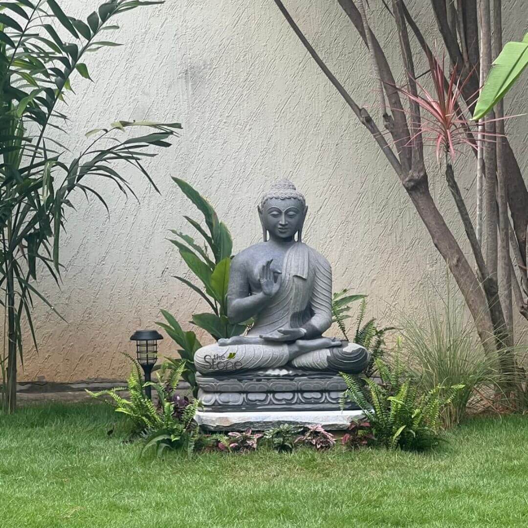 Black Granite Buddha Statue 2.5ft 3 A serene 2.5-foot grey granite Buddha statue is prominently displayed in a lush outdoor garden setting, embodying tranquility and natural harmony. The Buddha is seated in a cross-legged meditative pose, with his right hand raised in the Abhaya Mudra (gesture of fearlessness and reassurance) and his left hand resting gently in his lap. His eyes are downcast, and a subtle, peaceful smile adorns his face, conveying a sense of deep calm. The statue rests on a layered base, possibly a combination of carved stone and a simple white pedestal, elevated slightly above the vibrant green grass. Surrounding the base are various green plants, including ferns and broad-leafed foliage, which naturally integrate the statue into the landscape. To the left of the Buddha, a small modern solar garden light blends into the greenery. In the background, a light-colored stucco wall provides a neutral backdrop, while several slender tree trunks and the fronds of a palm tree frame the scene, enhancing the natural and contemplative atmosphere of this outdoor sacred space. The texture of the granite is visible, showcasing the durable and natural material suitable for outdoor placement.