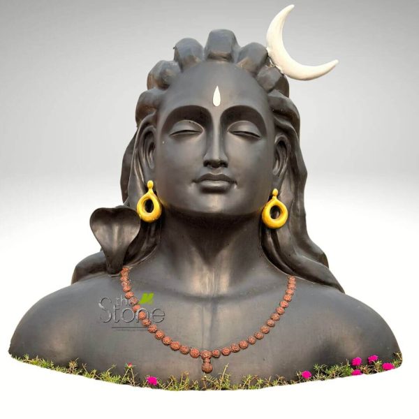 finely detailed, large black or dark grey stone bust depicting the Hindu deity Lord Shiva in a profound state of meditation, with his eyes gently closed and a calm, contemplative expression radiating tranquility. A distinctive white vertical tilak marks his forehead, symbolizing the third eye. His matted locks, or Jata, are elegantly styled, adorned with a brilliant white crescent moon (Chandra) placed on the right side of his head. He wears golden, teardrop-shaped earrings in both ears, and a traditional Rudraksha mala, made of numerous reddish-brown spherical beads with a central pendant, drapes gracefully around his neck and chest. A black cobra, likely Vasuki or Nagaraja, is artfully coiled around his left shoulder, its head subtly visible behind his ear. The sculpture is set against a simple, light grey background and emerges from a natural base of lush green plants interspersed with small, vibrant pink flowers, suggesting its placement as an exquisite garden statue or a focal point in a spiritual outdoor setting. This powerful and detailed artwork embodies tranquility and devotion, highlighting intricate elements of Hindu iconography and skilled craftsmanship, making it a perfect visual for articles discussing spiritual sculptures, Hindu traditions, garden decor, or artisanal stone works.