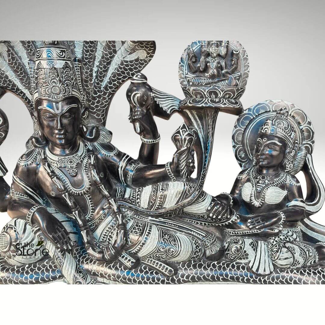 Lord Vishnu Sleeping on Sheshnag Statue 20" 6 magnificent and intricately carved dark gray stone sculpture, depicting the iconic "Anantashayana Vishnu" scene from Hindu mythology. Lord Vishnu is shown reclining serenely on the coiled body of the multi-headed cosmic serpent Sheshnaga (also known as Ananta). Vishnu, with a calm and divine expression, is adorned with an elaborate crown, numerous necklaces, armlets, bracelets, and anklets, all meticulously detailed. He is depicted with four arms: his upper left hand holds a disc (chakra), his upper right hand holds a conch shell (shankha), while his lower left hand rests on his thigh, and his lower right hand is extended forward. His garments are richly carved with intricate patterns, providing a beautiful contrast against the dark stone, highlighted by lighter, almost silvery etched details. Sheshnaga's body forms Vishnu's bed, with its multiple hooded heads (at least seven are clearly visible) fanning out majestically behind and above Vishnu's head, forming a protective canopy. The serpent's scales are also intricately textured. To Vishnu's right, at his feet, sits Goddess Lakshmi, his consort, elegantly adorned with her own crown and jewelry. She gazes towards Vishnu with devotion, her hands positioned near his feet, one possibly holding a small lotus bud. A beautiful, radiating circular halo is carved behind her head. Emerging from Vishnu's navel, a tall, ornate lotus stem gracefully rises upwards, culminating in a blooming lotus flower. Seated upon this lotus is the four-headed Lord Brahma, the creator god, in a meditative pose with multiple arms, also adorned with a crown and jewelry. The entire sculpture is a masterpiece of traditional Indian stone carving, where the dark, almost black, base material is contrasted with exquisitely fine white or light gray lines and patterns, bringing out every minute detail of the deities, their ornaments, and their surroundings. This detailed depiction symbolizes cosmic rest, creation, preservation, and the cyclical nature of time, making it a profound representation of divine cosmic order central to Hindu beliefs.