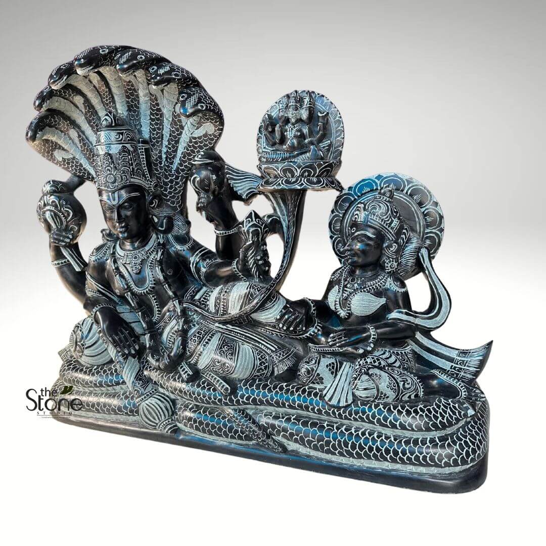 Lord Vishnu Sleeping on Sheshnag Statue 20" 5 detailed, dark gray or black stone sculpture depicts Lord Vishnu reclining on the cosmic serpent Sheshanaga, a central scene from Hindu mythology. Vishnu, portrayed with four arms, lies majestically on the coiled body of Sheshanaga. In his upper right hand, he holds a conch shell (shankha), and in his upper left, a discus (chakra). His lower left hand gently grasps a lotus stem that gracefully rises upwards, culminating in a seated figure of Lord Brahma atop the lotus flower. Vishnu is adorned with an elaborate, multi-tiered crown, necklaces, armbands, and a draped dhoti, all intricately carved with traditional Indian patterns that stand out in lighter tones against the dark stone. His face bears a serene and peaceful expression, characteristic of his divine slumber (Yoga Nidra). Sheshanaga forms a majestic bed and canopy for Vishnu. Its robust, scaled body is coiled to support him, while its seven cobra heads fan out dramatically behind Vishnu's head, providing a protective hood. Each serpent head and the body are meticulously etched with scales, eyes, and markings, creating a striking texture. To Vishnu's right, at his feet, sits Goddess Lakshmi, his consort, gazing devotedly towards him. She is depicted with two arms, her hands in a respectful or worshipful gesture, possibly massaging his feet. Lakshmi, too, wears an ornate crown, necklaces, earrings, and bangles, with intricate patterns adorning her garments. A circular, stylized halo (prabhamandala) frames her head, signifying her divine status. The entire sculpture rests on a flat base that is also carved with continuous scale patterns, visually extending the form of the cosmic serpent. The craftsmanship highlights the high skill in stone carving, utilizing contrasting textures and depths to bring out the mythical narrative. This traditional Indian art piece, beautifully lit against a plain white background, emphasizes the intricate details of the figures, their divine attributes, and the symbolism of cosmic creation and preservation embodied by Vishnu, Lakshmi, Sheshanaga, and Brahma. A small "The Stone Studio" watermark is visible in the bottom left corner.