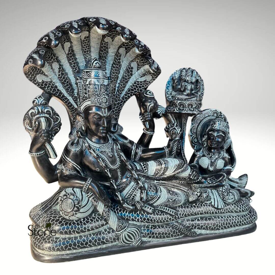 Lord Vishnu Sleeping on Sheshnag Statue 20" 4 Here's a long and descriptive alt text for the image: ```alt A beautifully carved, large black stone sculpture depicts the Hindu deity Lord Vishnu in his iconic Anantasayana pose, reclining on the coiled primordial serpent Shesha Naga, attended by his consort Lakshmi and Lord Brahma. The highly detailed carving features a striking contrast, with the dark, almost black stone forming the main figures and background, while intricate details are highlighted with a lighter, almost silvery grey finish, making them stand out. At the center, Lord Vishnu reclines serenely on the massive, coiled body of Shesha Naga, whose numerous cobra heads (approximately seven to nine are visible) fan out majestically above Vishnu like a protective canopy. Vishnu's face is calm and contemplative, his eyes partially open, signifying his state of cosmic sleep (Yoga Nidra). He is adorned with an elaborate, towering crown, intricate necklaces, armlets, bracelets, and anklets, all meticulously detailed. His four arms are gracefully positioned: the upper left hand holds a conch shell (shankha), the upper right holds a discus (chakra), the lower left hand holds a lotus flower (padma), and the lower right arm is bent at the elbow, resting on the serpent's coils, with the hand near his waist. His lower garment (dhoti) is also highly decorated with patterns. To Vishnu's right, seated gracefully near his feet, is his consort, Goddess Lakshmi. She is depicted with a serene expression, adorned with her own crown, necklaces, and bangles, her gaze directed towards Vishnu in devotion. Her right hand appears to be gently touching or massaging Vishnu's foot, a traditional gesture of service and reverence. Rising from the base between Vishnu and Lakshmi, on a separate ornate pillar-like structure, is a smaller representation of Lord Brahma, seated in meditation. He is depicted with multiple heads (three or four are visible), indicating his role as the creator god emerging from the lotus that typically sprouts from Vishnu's navel, symbolizing the start of a new cosmic cycle. The entire sculpture rests on a solid, dark stone base. The craftsmanship is exceptional, evident in the delicate scales of the serpent, the individual folds of the deities' garments, the intricate patterns of their jewelry, and their expressive facial features. This black stone idol perfectly encapsulates the mythological narrative of Vishnu as the preserver of the universe, resting between cycles of creation and dissolution, a significant piece of traditional Hindu religious art and iconography. ```
