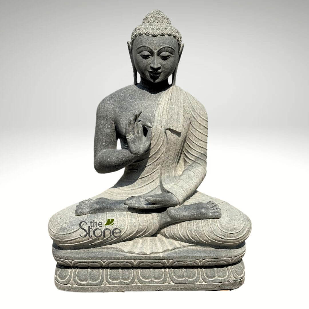 Black Granite Buddha Statue 2.5ft 1 full-length, front-facing view of a serene, hand-carved Buddha statue made from dark grey, finely speckled natural stone, possibly granite or basalt. The statue depicts Siddhartha Gautama seated in the meditative Padmasana (lotus) position, with his legs fully crossed, feet resting on opposing thighs, symbolizing profound stability and enlightenment. Buddha's right hand is raised to shoulder height with the palm facing outwards, fingers extended upwards and touching at the tips in the Abhaya Mudra, a traditional gesture of fearlessness, reassurance, and divine protection. His left hand rests gently in his lap, palm facing upwards, signifying contemplation, receptivity, and generosity. His face conveys deep tranquility and inner peace, with eyes half-closed in a state of introspection, a gentle, subtle smile, and elongated earlobes. His hair is meticulously depicted as tightly coiled curls or snail shells, topped by the Usnisha, the traditional cranial protuberance signifying wisdom and spiritual attainment. The flowing monastic robes are artfully draped over his left shoulder, leaving the right shoulder exposed, with realistic folds and creases that add depth and movement to the static form, showcasing the artisan's skill in stone carving. The figure rests upon a sturdy, multi-tiered rectangular pedestal. The lower tier features a carved decorative border of traditional lotus petals, while the upper tier is smooth, providing a stable foundation for the meditative posture. The statue is presented against a plain, light grey background, highlighting its intricate details and spiritual presence. This finely crafted artwork emphasizes spiritual calm, mindfulness, and the timeless principles of Buddhism, making it an ideal piece for meditation spaces, gardens, or as a focal point in sacred home decor, showcasing the enduring beauty of natural stone sculpture.