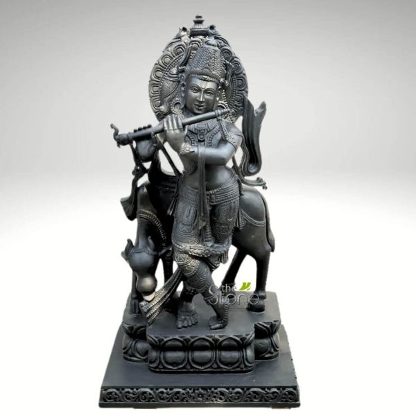 magnificent, intricately carved dark stone or bronze-like sculpture depicts Lord Krishna standing gracefully in a classic tribhanga (three-bend) pose, playing his bansuri (flute). His serene face features a gentle smile, radiating peace and divine charm. He wears an elaborate, tiered crown adorned with detailed patterns, likely including a peacock feather motif, which signifies his divine majesty. Krishna is richly adorned with finely sculpted traditional jewelry, including layered necklaces, armlets, wristbands, and anklets, alongside a beautifully draped dhoti-like garment around his lower body, showcasing intricate folds and patterns. Behind him, a majestic cow stands, its head gently turned towards Krishna, symbolizing his role as Gopala (the divine cowherd) and his deep connection to nature and animals. The head of another cow is subtly visible near his left leg, further emphasizing this pastoral theme. A large, semi-circular prabhamandala (halo or aureole) with ornate carvings, possibly depicting floral or geometric patterns, frames his upper body and head, signifying his divinity. The entire sculpture rests on a multi-tiered rectangular base, which is also decorated with traditional Hindu motifs such as lotus petals and classical architectural patterns. The dark, monochromatic finish of the sculpture highlights the exceptional craftsmanship, the intricate details, and the varied textures, making it a striking piece of traditional Indian art. This artwork beautifully encapsulates themes of devotion, harmony, divine music, and Hindu mythology, making it highly relevant for articles discussing spiritual artifacts, traditional Indian craftsmanship, devotional items, or home decor inspired by cultural heritage.