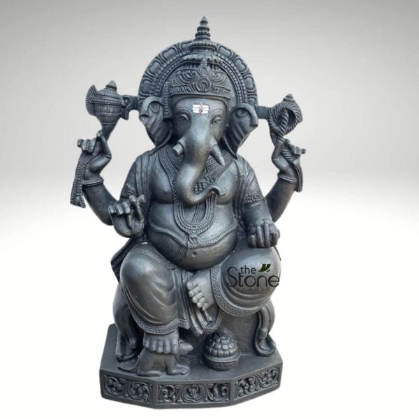 majestic and intricately carved dark gray stone statue of Lord Ganesha, the revered Hindu deity, is presented against a plain, light gray studio background. This handcrafted sculpture captures Ganesha in a traditional seated posture, radiating a sense of calm and wisdom. Ganesha, with his iconic elephant head, features prominent, large ears, serene half-closed eyes, and a distinct white and red tilak mark on his forehead. His trunk curls gently to his left, gracefully reaching towards a small bowl of modaks (sweets) held in his lower left hand, symbolizing his love for offerings and abundance. He wears an ornate, tiered crown and a grand circular prabhamandala (halo) richly decorated with radial and scrollwork carvings behind his head, signifying his divine presence and aura. His four arms are gracefully depicted, each holding symbolic items. In his upper right hand, he holds an ankusha (elephant goad), and in his upper left, a pasha (noose), representing control over worldly attachments and obstacles. His lower right hand is raised in the abhaya mudra, a gesture of blessing, reassurance, and dispelling fear for devotees. The pot-bellied Ganesha is adorned with an array of intricate jewelry, including multiple necklaces, detailed armlets, wristlets, and a sacred thread (yagnopaveetam) draped across his chest, all sculpted with fine detail. He is seated comfortably on an ornate, multi-layered pedestal, wearing traditionally draped garments. At his left foot (our right), his faithful vahana (mount), Mooshika (a small mouse), sits attentively, symbolizing humility and Ganesha's ability to overcome even the smallest obstacles. A small bowl or pot, possibly containing more modaks, rests beside Mooshika. The sturdy rectangular base itself is beautifully embellished with classical Indian scrollwork and floral motifs, adding to the statue's grandeur and stability. The statue appears to be crafted from high-quality dark stone, possibly granite or a similar material, with a smooth, polished finish that expertly highlights the depth and precision of the carvings. The overall impression is one of exquisite craftsmanship and spiritual elegance, making this Ganesha idol an ideal centerpiece for a Hindu altar, puja room, or as a sophisticated piece of spiritual home decor. A "The Stone Studio" watermark with a subtle leaf icon is visible on the lower right, identifying the artisan or manufacturer of this divine Ganesha sculpture, suggesting its availability as a premium art or spiritual item.