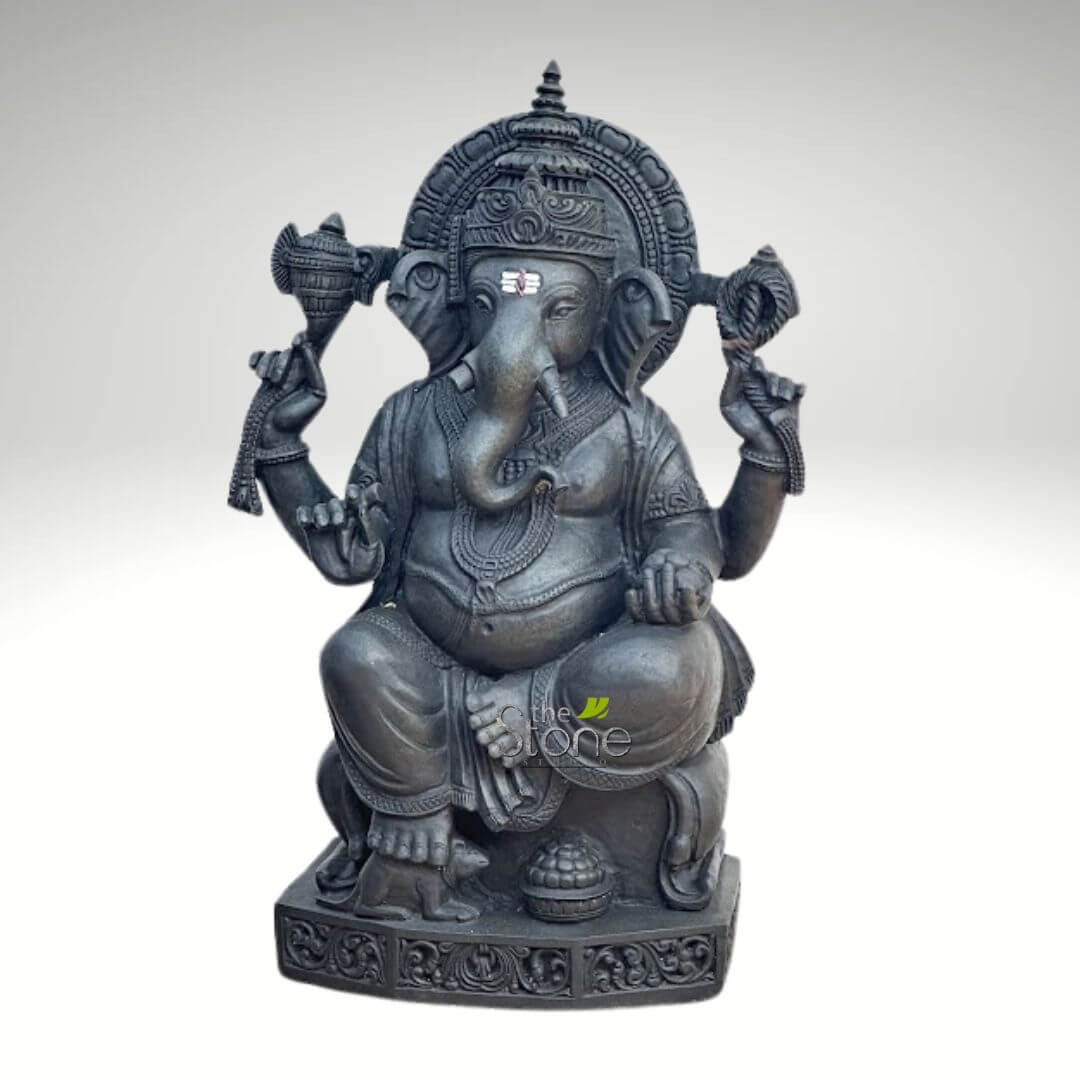 Ganapathi Statue for Home 2.5ft 5 "A meticulously crafted, dark-toned stone sculpture of Lord Ganesha, seated serenely against a plain light grey background. The idol, likely carved from black granite or basalt, showcases exceptional detail and a matte finish. Ganesha is depicted with his iconic elephant head, featuring large, expressive ears, small eyes, and a gently curving trunk that turns left towards his lower left hand. A prominent red and white tilak (sacred mark) adorns his forehead, and one tusk appears stylized or broken, a traditional characteristic. He possesses four arms, symbolizing his divine power and various attributes. In his upper right hand, Ganesha holds an ankusha (elephant goad), while his upper left hand grasps a pasha (noose). His lower right hand delicately holds a japamala (prayer beads), and his lower left hand extends forward, offering or holding a modak (sweet dumpling), towards which his trunk is poised. Ganesha's portly, pot-bellied form, a symbol of prosperity and abundance, is adorned with intricate jewelry, including necklaces, armlets, and anklets, along with a beautifully draped lower garment. He sits comfortably on an elaborately carved pedestal. At his feet, to the left, is a small, loyal mouse (mooshika), his traditional vehicle, and beside it, a small bowl overflowing with modaks, signifying offerings and blessings. Behind his head, a magnificently sculpted prabhamandala (aura or halo) with ornate patterns frames the deity, adding to his grandeur. This exquisite Ganesha idol is a powerful representation suitable for articles exploring traditional Indian stone carving, Hindu religious art, spiritual home decor, the significance of Lord Ganesha as the remover of obstacles and bestower of wisdom and good fortune, or high-quality artisanal sculptures for worship and aesthetic appeal."