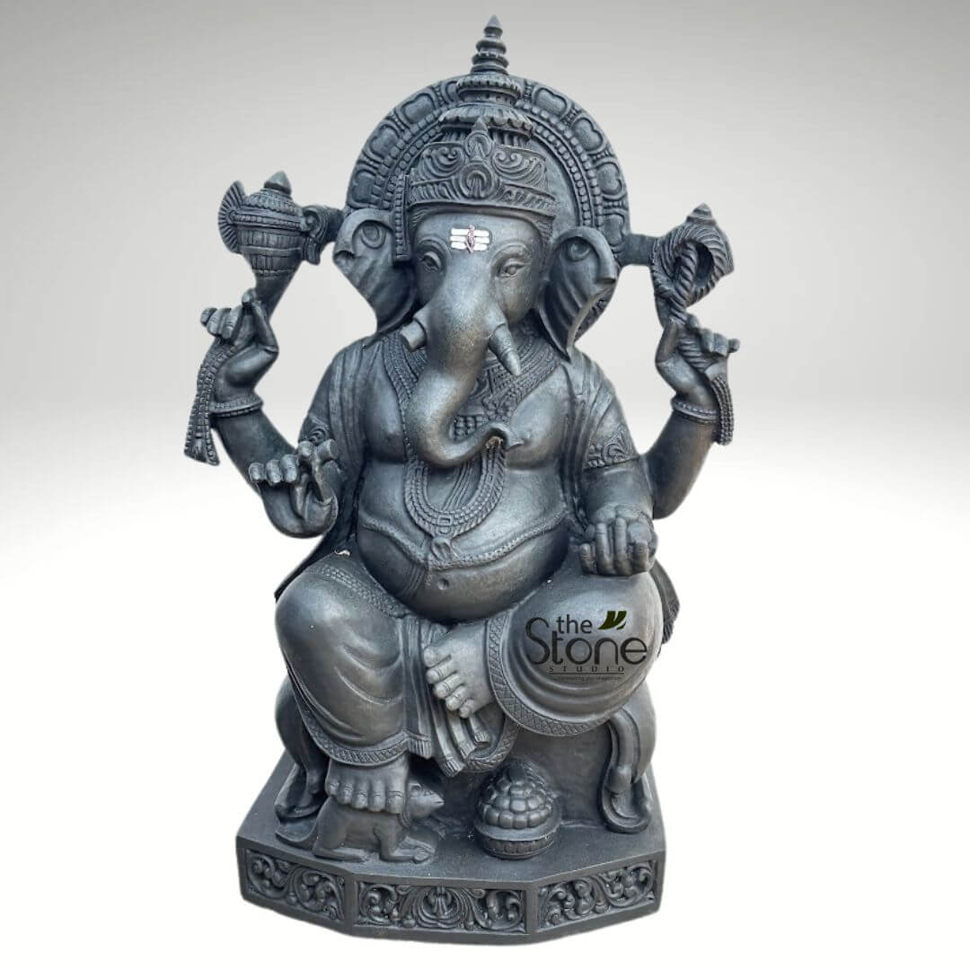 Ganapathi Statue for Home 2.5ft 4 meticulously carved, dark grey stone statue of Lord Ganesha, seated in a traditional posture, is presented against a plain, light grey background. The idol's rich, almost charcoal-black hue, possibly polished granite or black marble, imparts a sense of gravity and timelessness. Ganesha, the popular Hindu deity revered as the remover of obstacles and god of wisdom and prosperity, sits comfortably with his legs folded, one foot subtly raised, projecting an aura of serenity and grace. His iconic elephant head is finely sculpted, featuring large, expressive ears, a prominent trunk curling gracefully towards his left (viewer's right), and a distinctive single tusk (Ekadanta Ganesha). A small, yet vibrant white and red tilaka mark adorns his forehead, symbolizing auspiciousness and spiritual awakening. He wears an ornate, multi-tiered crown (mukuta) with a spire at the top, and is backed by an intricately carved prabhavali (arch or halo), both adding to his majestic presence. The statue features four arms, each holding symbolic attributes. In his upper right hand, he holds an ankusha (elephant goad), signifying mastery over desires, while his upper left hand grasps a pasha (noose), symbolizing spiritual attachment and restraint. His lower right hand rests gracefully on his knee, holding a sweet modak, while his lower left hand cradles a large bowl brimming with laddoos or modaks, reflecting his love for sweets and association with abundance. Ganesha is adorned with elaborate necklaces, armlets, wristlets, and a waistband, all meticulously carved into the stone, highlighting the traditional craftsmanship. His round belly, a symbol of prosperity and the ability to digest all experiences, is also prominently depicted. At his feet, to his left, sits a small, alert mouse (Mooshika), Ganesha's loyal vahana (vehicle), symbolizing the conquering of desire and ego. The entire idol rests upon a robust, rectangular base, decorated with intricate floral and geometric relief carvings, providing a sturdy and aesthetically pleasing foundation. The 'the Stone STUDIO' watermark in white text is subtly visible in the lower right quadrant, identifying the creator or retailer and emphasizing that this is a premium, handcrafted religious statue made from natural stone. This detailed Lord Ganesha murti is an exquisite example of Hindu deity art, perfect for home altars, meditation spaces, temple decor, or as a significant cultural and spiritual art piece. Its durable material and intricate stone carving make it a timeless representation of spiritual wisdom and prosperity.