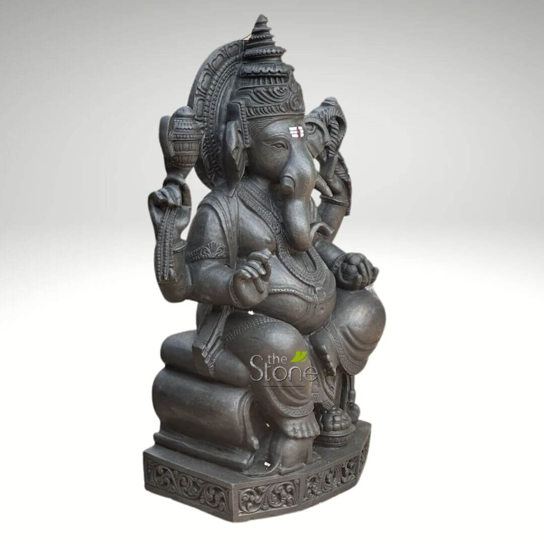 Ganapathi Statue for Home 2.5ft 3 meticulously crafted, dark gray stone statue of Lord Ganesha, the revered Hindu elephant-headed deity, is depicted in a full-body view against a plain, light gray background. The sculpture, likely made from black granite or a similar natural stone, showcases intricate detail and traditional Indian artistry. Ganesha is seated majestically on an ornate, multi-tiered pedestal, which itself rests upon a rectangular base adorned with continuous, symmetrical scrollwork and traditional patterns. His plump form, a symbol of prosperity and abundance, is richly adorned with finely carved traditional jewelry, including necklaces, armlets, wristlets, anklets, and a prominent snake-shaped belt around his belly. He possesses four arms: his upper left hand holds a pot or vessel, possibly filled with modakas or other auspicious offerings; his upper right hand grasps an ankusha (elephant goad); his lower left hand is extended outwards, palm facing upwards in a gesture of blessing (varada mudra); and his lower right hand delicately holds a modaka (sweet dumpling), towards which his trunk gracefully curves to the left, symbolizing his love for sweets and accessibility to his devotees. Ganesha's benevolent elephant head features large, fanned ears and a single, intact tusk, signifying his Ekadanta form. A distinct red and white tripundra tilak is prominently marked on his forehead, representing spiritual devotion. He wears an elaborate, tiered crown (mukuta) with finely detailed carvings, and behind his head, an intricately sculpted prabhavali (decorative arch or aura) showcases traditional motifs, enhancing the statue's divine presence. The texture of the stone is visible, adding to the authentic, handcrafted appeal of this sacred idol, suitable for spiritual decor, puja rooms, or as a timeless piece of Hindu art.