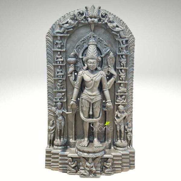 tall, monochromatic dark grey stone sculpture depicts a four-armed Hindu deity, almost certainly Lord Vishnu, standing centrally in an intricate bas-relief. The deity is adorned with a tall, ornate crown (kiritamukuta) and elaborate jewelry including necklaces, armlets, bracelets, and a waist belt, all finely detailed. Vishnu's front right hand holds a lotus bud, his front left hand rests on a club or mace (gada), his upper left hand grasps a conch shell (shankha), and his upper right hand holds a discus (chakra). He stands in a serene posture on a multi-tiered, circular pedestal, itself resting on a broader rectangular base. The central figure is framed by an ornate, arch-shaped prabhavali (aureole or archway). This archway is richly decorated with a series of smaller narrative panels. Along the inner edges of the arch, there are ten distinct rectangular niches on each side, vertically stacked, likely depicting the Dashavatara (ten incarnations of Vishnu), such as Matsya (fish), Kurma (tortoise), Varaha (boar), Narasimha (man-lion), and Vamana (dwarf), followed by human forms like Parashurama, Rama, Balarama or Krishna, Buddha, and Kalki. The apex of the arch features a prominent Kirtimukha (glory-face) motif, with celestial beings or flying figures in relief on the curved upper sections, flanked by decorative scrolls. Two larger, standing attendant figures, possibly consorts or celestial beings, are carved in relief at the base of the archway, flanking Vishnu. At the very bottom of the sculpture, on the pedestal beneath these attendants, additional smaller figures, appearing as devotees or sages, are depicted kneeling in adoration. The entire piece showcases exceptional traditional Indian stone carving and craftsmanship, with every element meticulously detailed, emphasizing mythological themes and religious iconography. The dark grey, matte finish of the stone evokes a sense of ancient artistry and spiritual gravitas. A transparent watermark for "the Stone" is subtly visible on the lower right side of the sculpture. This sculpture would be highly relevant to articles discussing Hindu deities, traditional Indian temple art, the Dashavatara concept, or the history and techniques of stone carving in India.