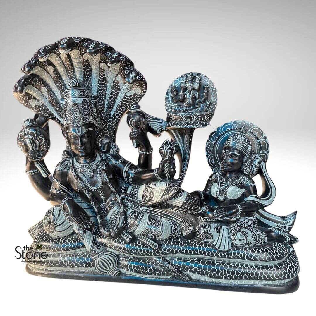 Lord Vishnu Sleeping on Sheshnag Statue 20" 3 magnificent, intricately carved sculpture crafted from dark, possibly black or dark gray soapstone, with exquisite details highlighted by a lighter, off-white finish. The central focus is a depiction of Lord Vishnu, the preserver god, reclining serenely on the coiled body of the multi-headed cosmic serpent, Sheshanaga (also known as Ananta). Vishnu is shown with a semi-upright torso, gazing forward with a calm, meditative expression. He wears an elaborate, tiered crown and is richly adorned with intricate necklaces, armlets, bracelets, and a patterned waist belt. He possesses four arms: his upper right hand holds the chakra (discus), his upper left grasps the shankha (conch shell), his lower left hand rests on his thigh holding a gada (mace/club), and his lower right hand delicately holds a lotus bud. His lower garment (dhoti) is meticulously sculpted with detailed patterns, including waves and geometric designs. Behind Vishnu's head, the multiple, possibly seven or nine, cobra heads of Sheshanaga fan out, forming a protective and majestic canopy. Each cobra head is distinct, with engraved scales and eyes. The serpent's colossal body forms Vishnu's celestial bed, its numerous coils detailed with an overlapping scale pattern that extends across the entire base of the sculpture, creating a textured and impressive foundation. To Vishnu's right, seated gracefully on the serpent's coils and facing him with an expression of devotion and attentiveness, is his consort, Goddess Lakshmi, the goddess of wealth and prosperity. She is adorned with a delicate crown, earrings, necklaces, and armlets, and her sari-like garment also features fine carved patterns. Her right hand rests affectionately on Vishnu's leg or foot, while her left hand holds a small lotus bud near her chest, symbolizing purity and beauty. Emerging from Vishnu's navel is a tall, elegant lotus stalk, culminating in a blooming lotus flower. Upon this lotus, Lord Brahma, the creator god, is seated in a meditative posture. Brahma is depicted with his characteristic four heads, symbolizing his omnipresence in the act of creation. The stark contrast between the dark stone and the fine, light-colored carved lines makes every detail pop, from the individual scales of the serpent to the deities' elaborate jewelry, garments, and facial features. This sculpture beautifully encapsulates the Hindu concept of Yoganidra (cosmic sleep) and the cyclical nature of creation, preservation, and dissolution, making it a significant representation of Vaishnava iconography and a testament to intricate stone carving artistry. It serves as a visual narrative of divine cosmic order and the roles of key deities in Hindu mythology. The "The Stone" watermark is visible in the lower left corner.