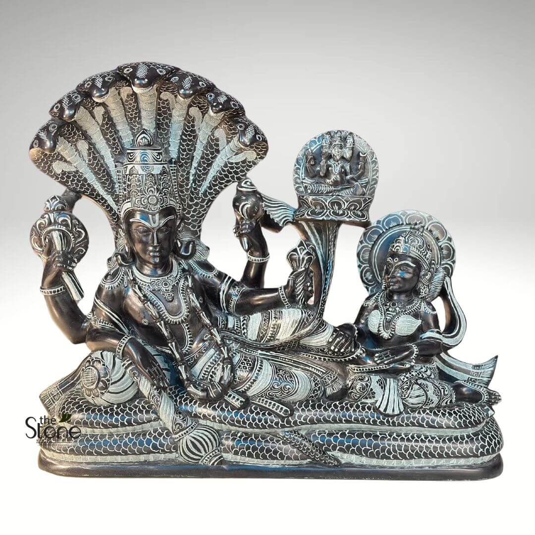 Lord Vishnu Sleeping on Sheshnag Statue 20" 1 meticulously carved, dark stone sculpture depicts the Hindu deity Lord Vishnu in his iconic cosmic slumber, known as Anantashayana, resting upon the multi-headed serpent Shesha. Lord Vishnu, centrally positioned, is shown reclining gracefully with his left arm bent, supporting his head. He possesses four arms, each holding one of his significant attributes: the Shankha (conch shell) in his lower left hand, the Chakra (discus) in his upper right, the Gada (mace) in his upper left, and a blossoming lotus stem emanating from his lower right hand, symbolizing creation. His serene and powerful countenance is framed by an elaborate, multi-tiered crown, and his body is adorned with richly detailed necklaces, armlets, bracelets, and a waist belt. His dhoti is intricately patterned with delicate, etched lines, showcasing traditional Indian craftsmanship. The colossal figure of Shesha-naga (or Ananta) forms both Vishnu's celestial bed, with its tightly coiled body providing a stable base, and a majestic canopy, as its numerous hooded heads fan out protectively behind Vishnu's head. Each serpent head is distinct, and the scales across Shesha's body are meticulously carved with fine, contrasting lines, creating a remarkable texture. Beside Vishnu, his divine consort, Goddess Lakshmi, is exquisitely carved, seated gracefully with a devoted expression. She gazes towards Vishnu, her hands in a respectful gesture, embodying loyalty and prosperity. Lakshmi is also adorned with a magnificent crown, intricate jewelry, and a flowing sari, all rendered with similar delicate etched patterns. Emerging from a lotus flower that blossoms from a stem originating from Vishnu's navel, Lord Brahma, the creator deity, is depicted seated. Though smaller in scale, his multi-headed form (with three heads visible from this perspective) is clearly discernible atop the lotus, signifying the cyclical nature of creation in Hindu cosmology. The entire sculpture is crafted from a dark, polished stone, possibly black granite or soapstone. The intricate details of the deities' clothing, ornaments, facial features, and the serpent's scales are brought to life through precise, light-colored linear etchings that contrast starkly against the dark background, giving the piece profound visual depth and highlighting the artistry. This detailed representation of the Vishnu Anantashayana scene serves as a powerful visual narrative for discussions on Hindu mythology, divine iconography, and traditional stone carving techniques.